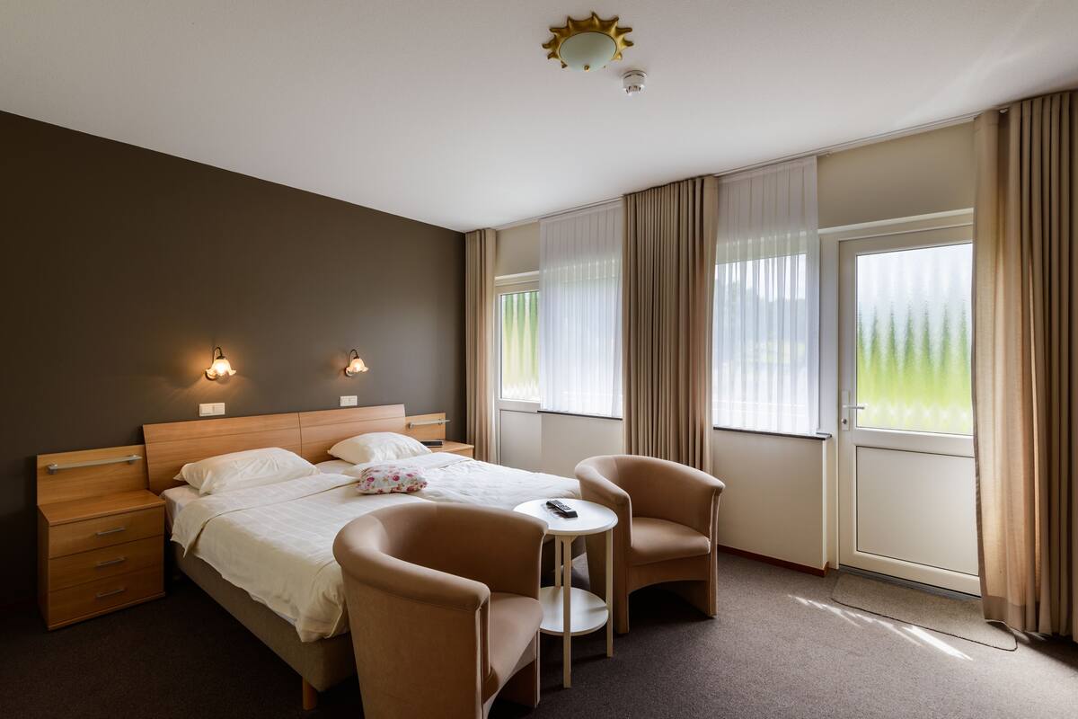 A hotel room with a bed, two chairs, a small table, and a window with curtains.