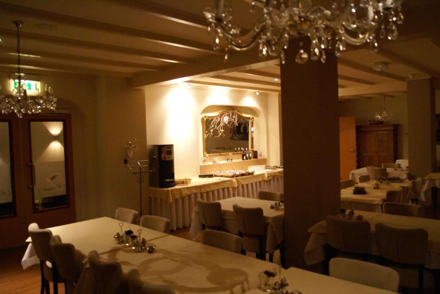 Un comedor con varias mesas y sillas, una zona de bar con un espejo y taburetes, y una araña.