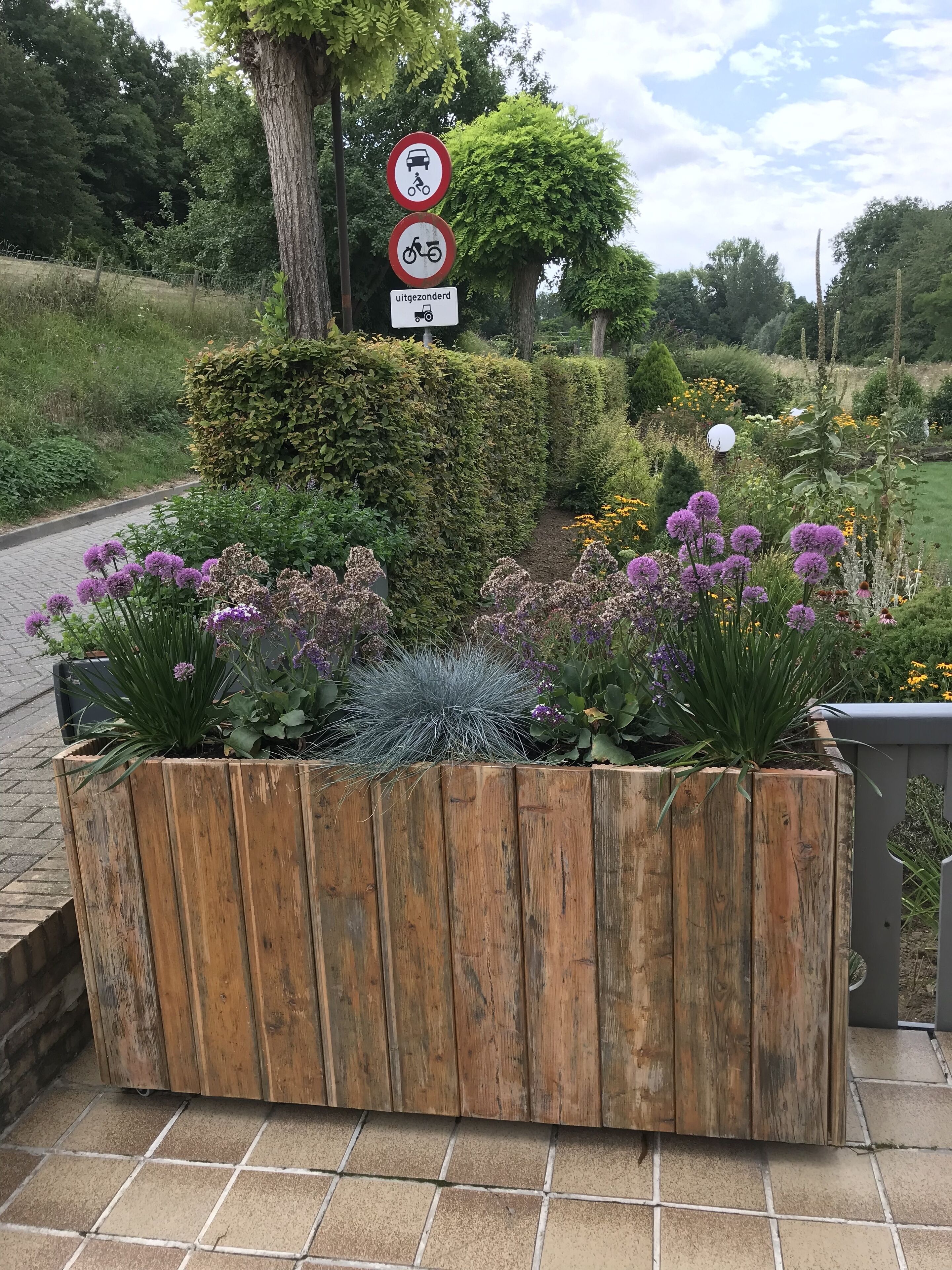 Een houten plantenbak met diverse planten en bloemen, een verbodsbord voor voertuigen en fietsen, en een bord dat aangeeft dat het gebied verboden is.