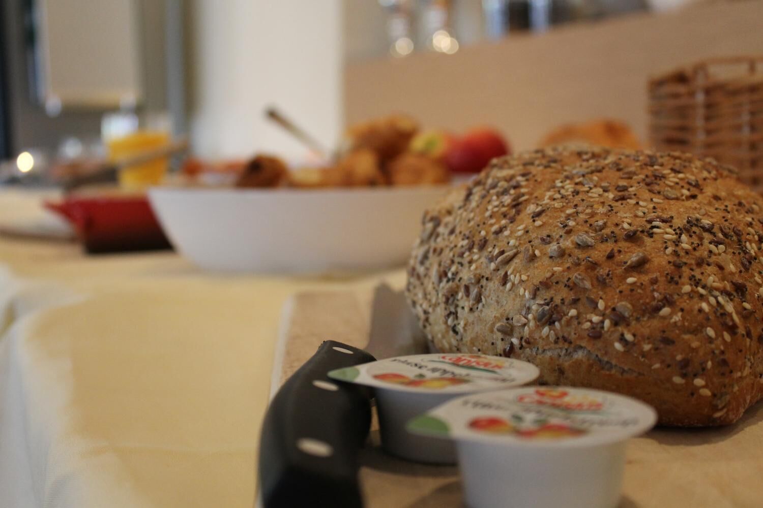 Una hogaza de pan con semillas sobre una mesa, junto a un cuchillo y dos recipientes de yogur.