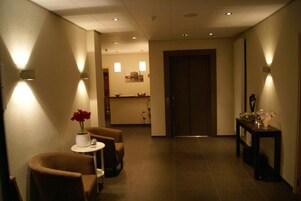 Um hall de hotel com balcão de recepção, área de estar com duas cadeiras e uma pequena mesa, e uma porta que dá para outro espaço.