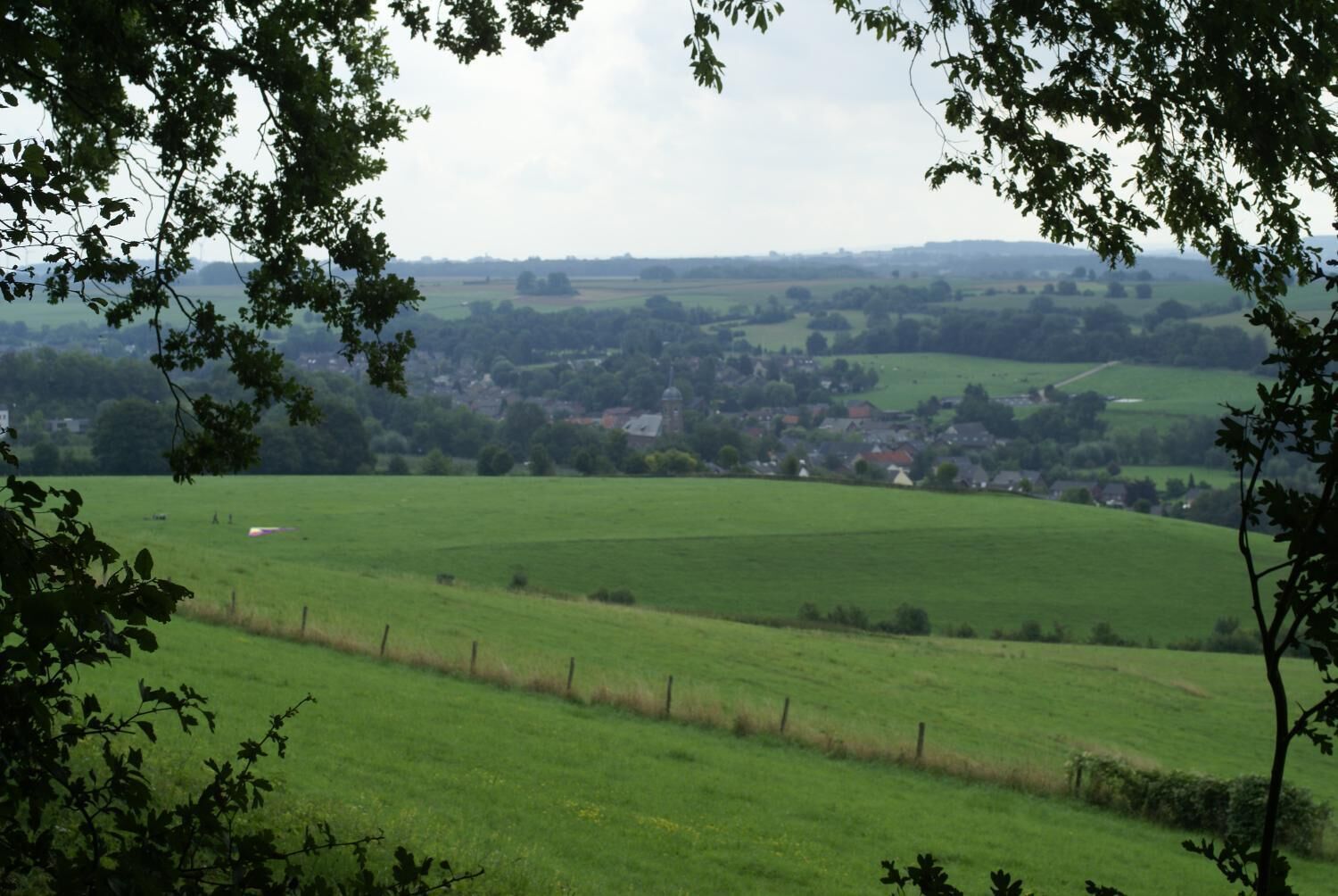 Een landelijk landschap met een groen veld, een omheining en een dorp in de verte.