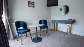 A room with two blue chairs around a small round table, a vanity with a mirror, and a television mounted on the wall.