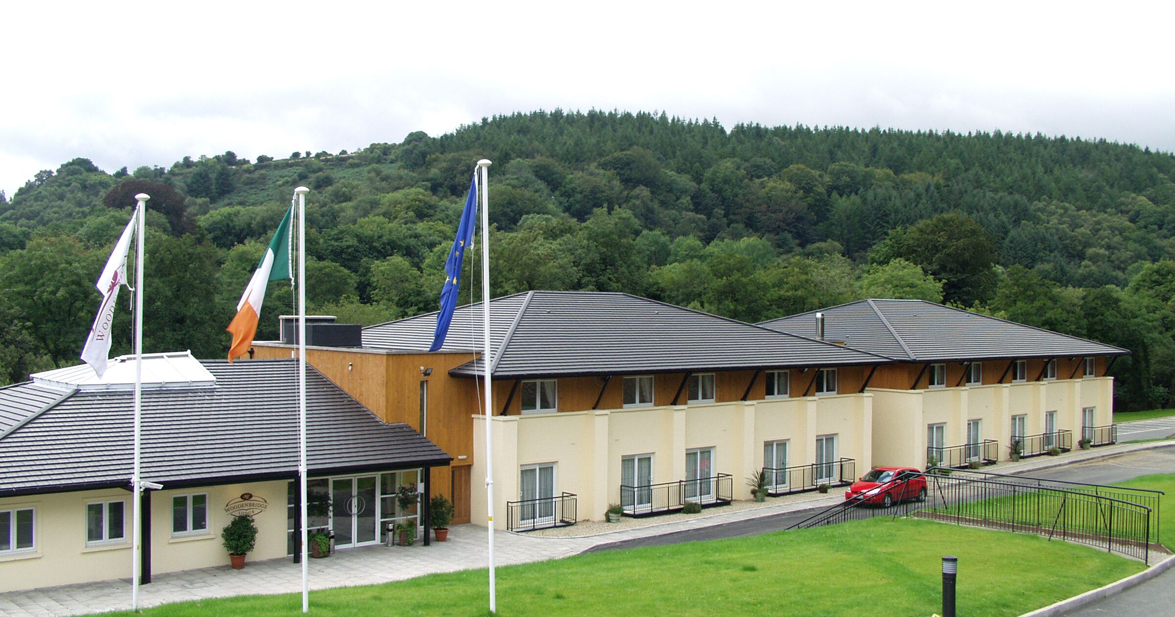 Een hotel met modern design, met een plat dak en een combinatie van beige en bruine gevels. Er zijn meerdere vlaggen, waaronder de Ierse vlag, geplaatst op vlaggenmasten. Het hotel ligt in een groen, heuvelachtig gebied met een weg ervoor.