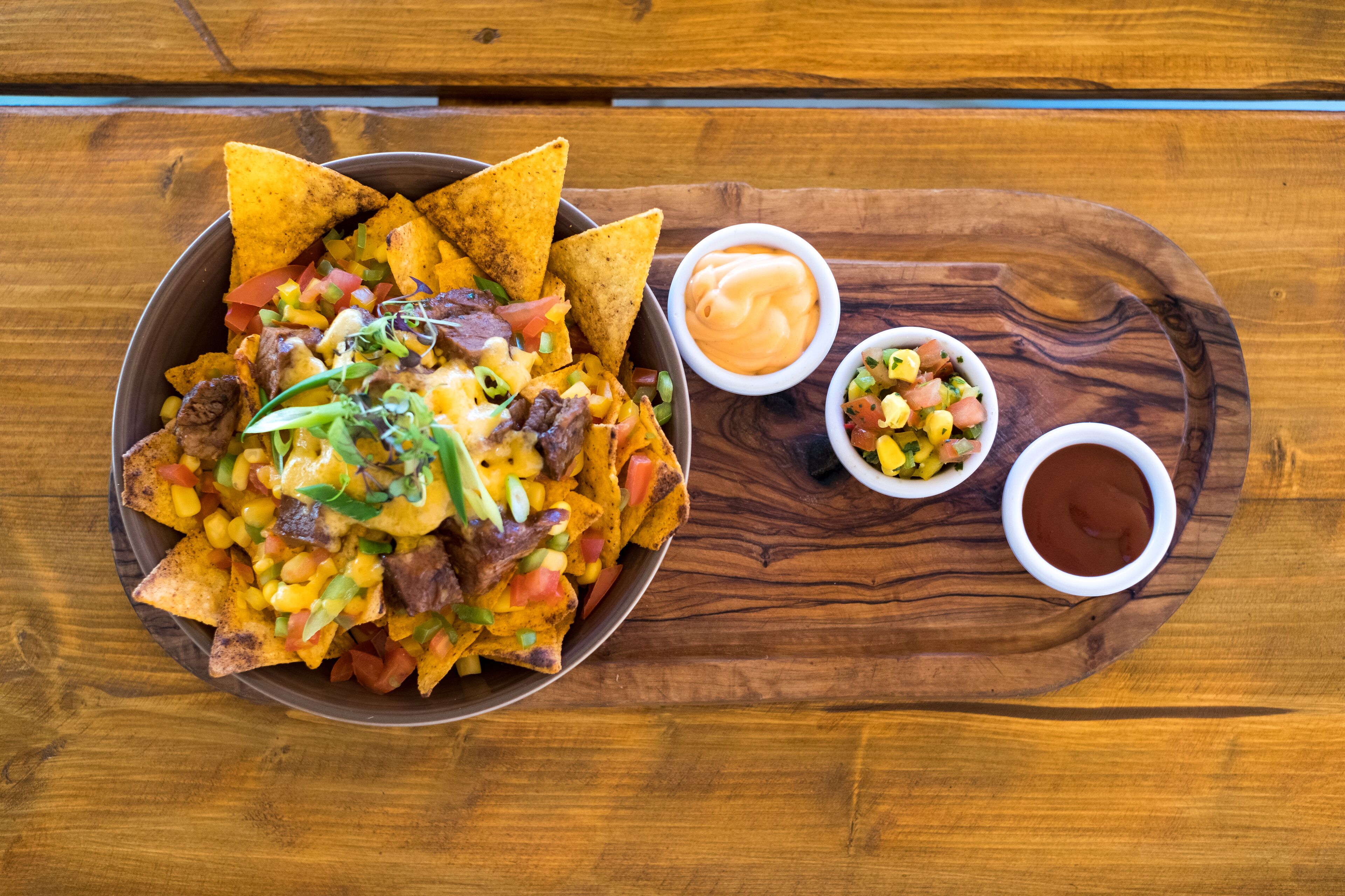 A bowl of nachos with toppings and two small bowls of dipping sauces on a wooden tray.