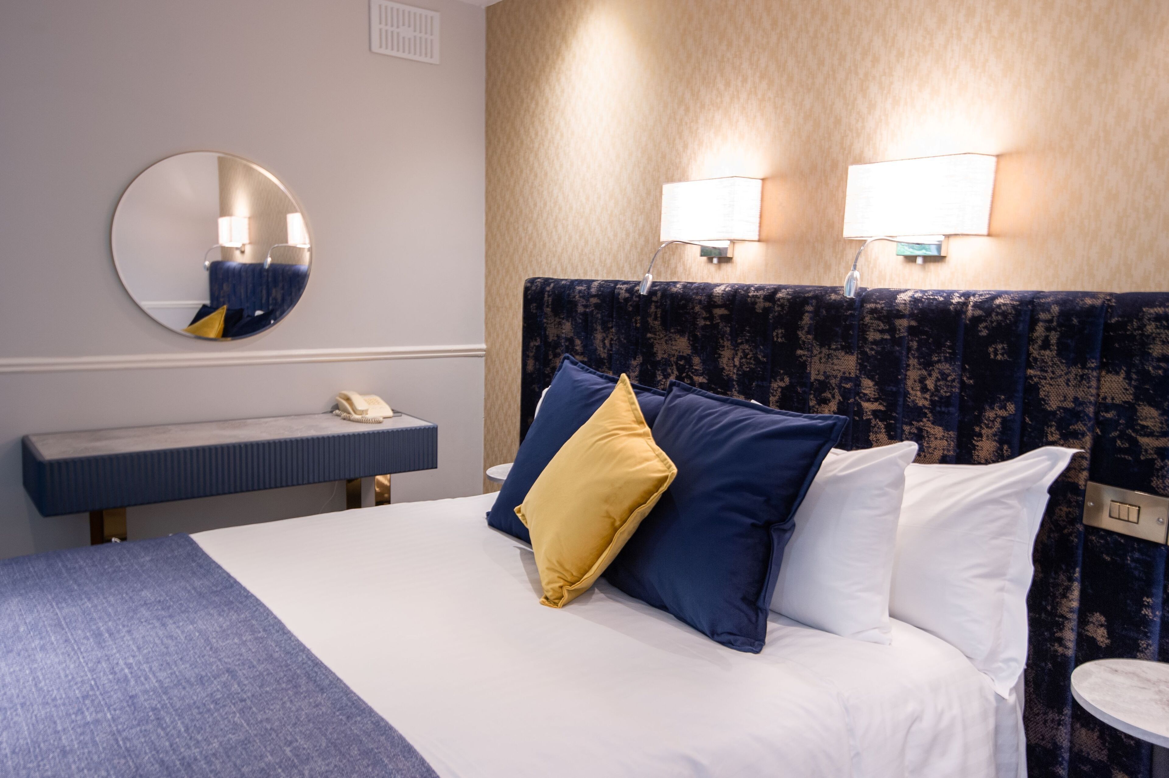 A hotel room with a bed, a round mirror, a bedside table with a phone, and two wall-mounted lamps.