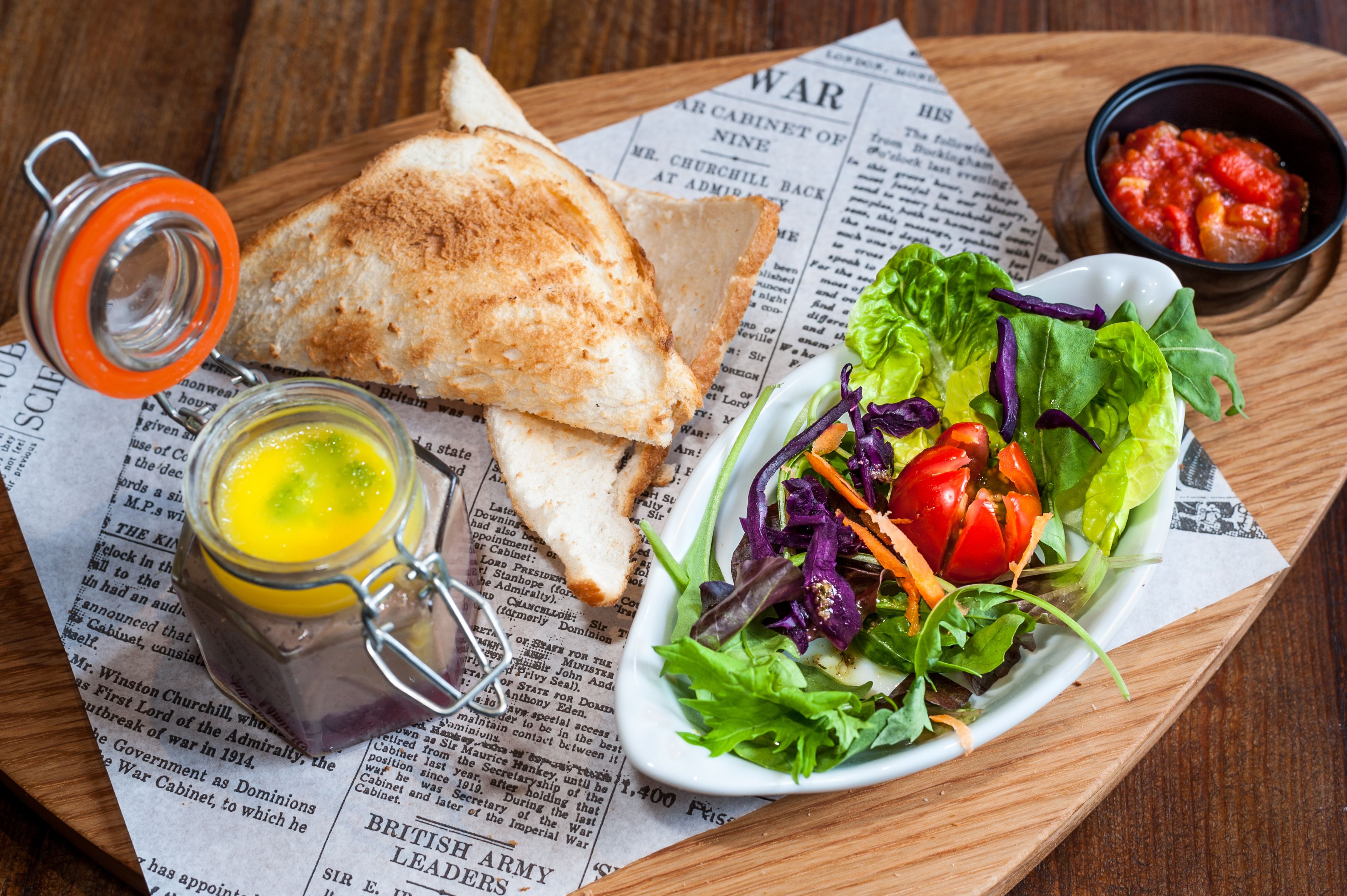 A sandwich on a newspaper, a salad, and a small jar of mustard.