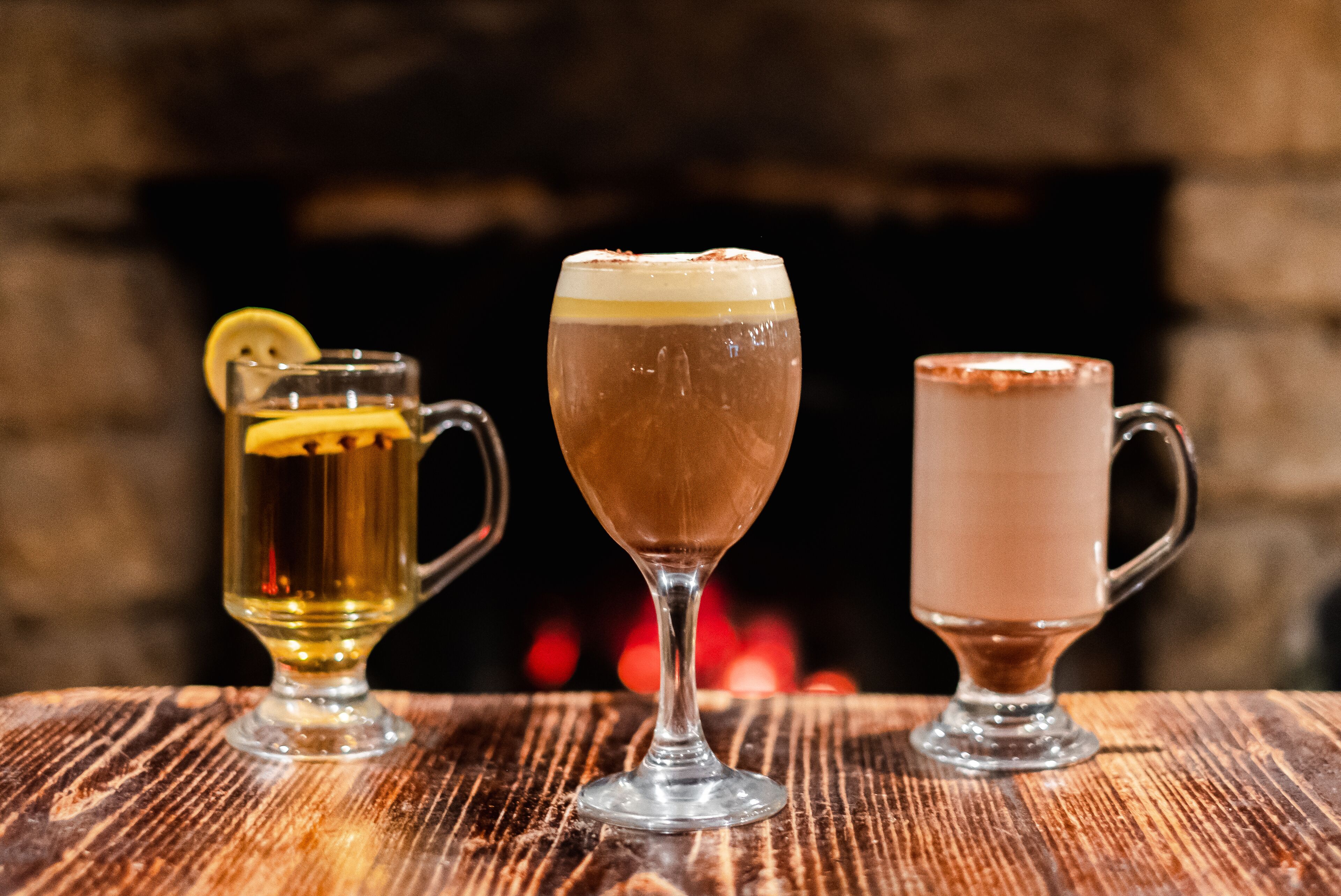 Tres vasos de bebidas sobre una superficie de madera con una chimenea al fondo.