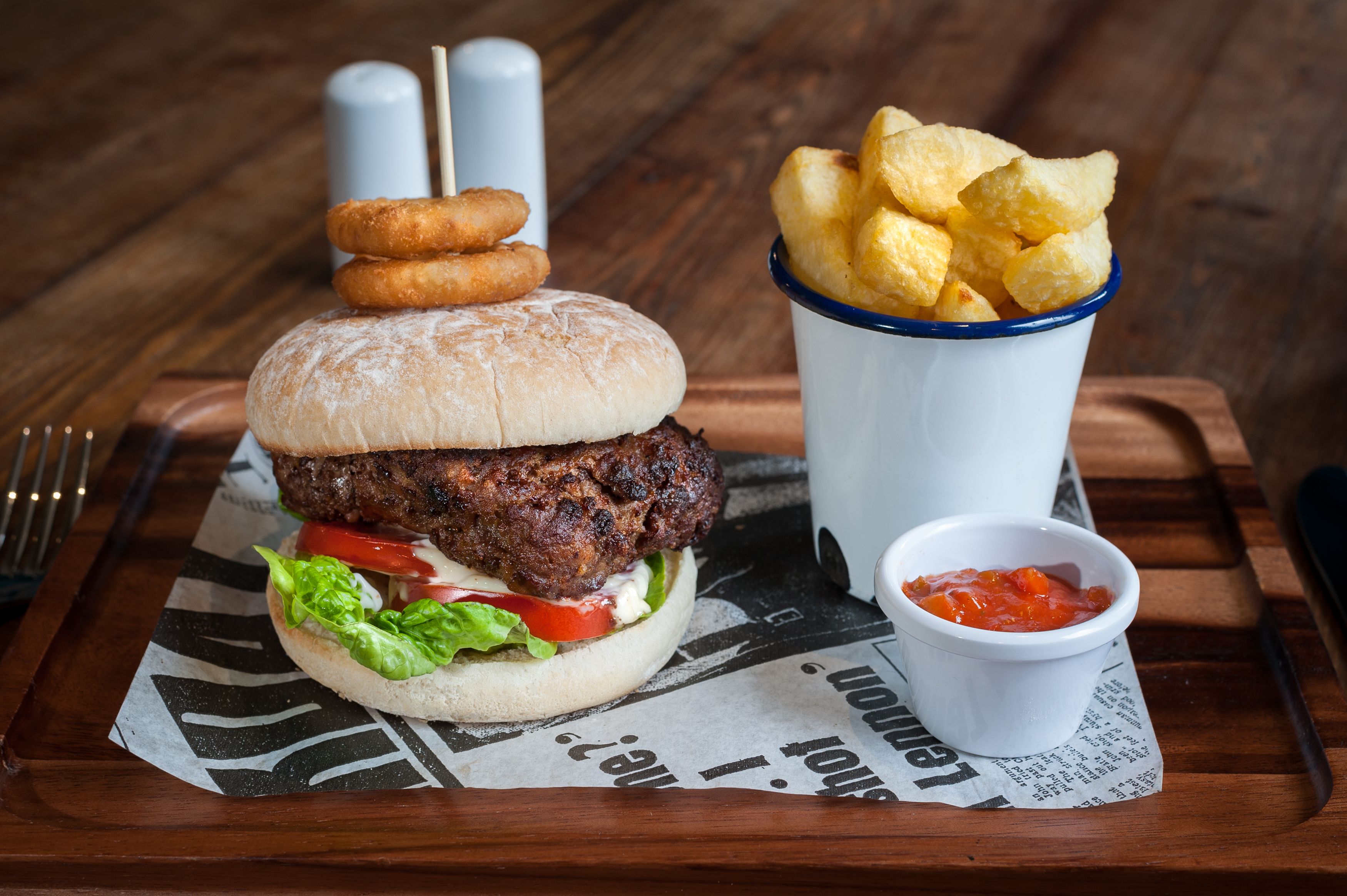 A burger with a side of chips and a small cup of sauce on a wooden table.