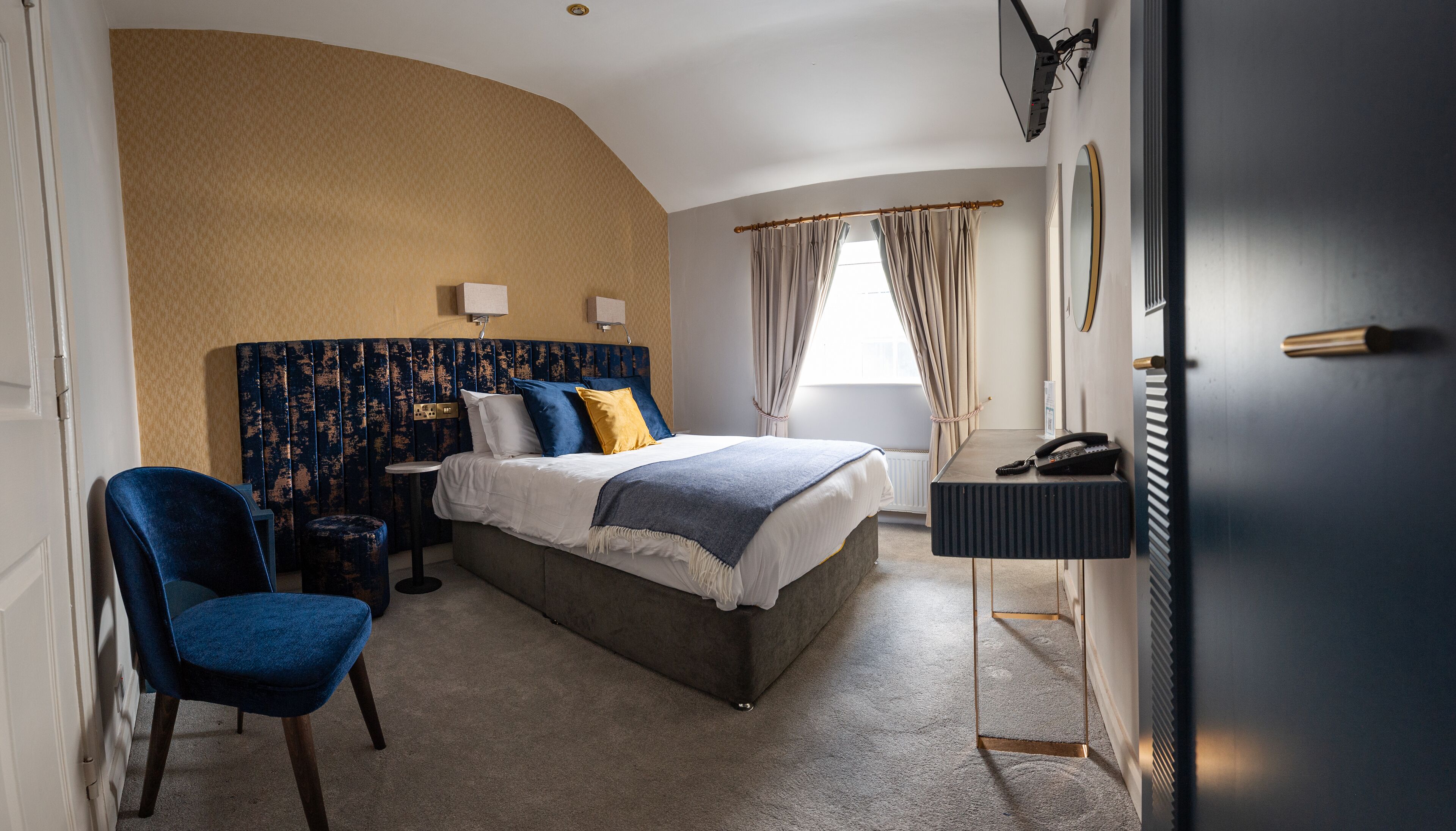 A bedroom with a curved wall, a bed with a headboard, a bedside table, a chair, a desk with a telephone, and a window with curtains.