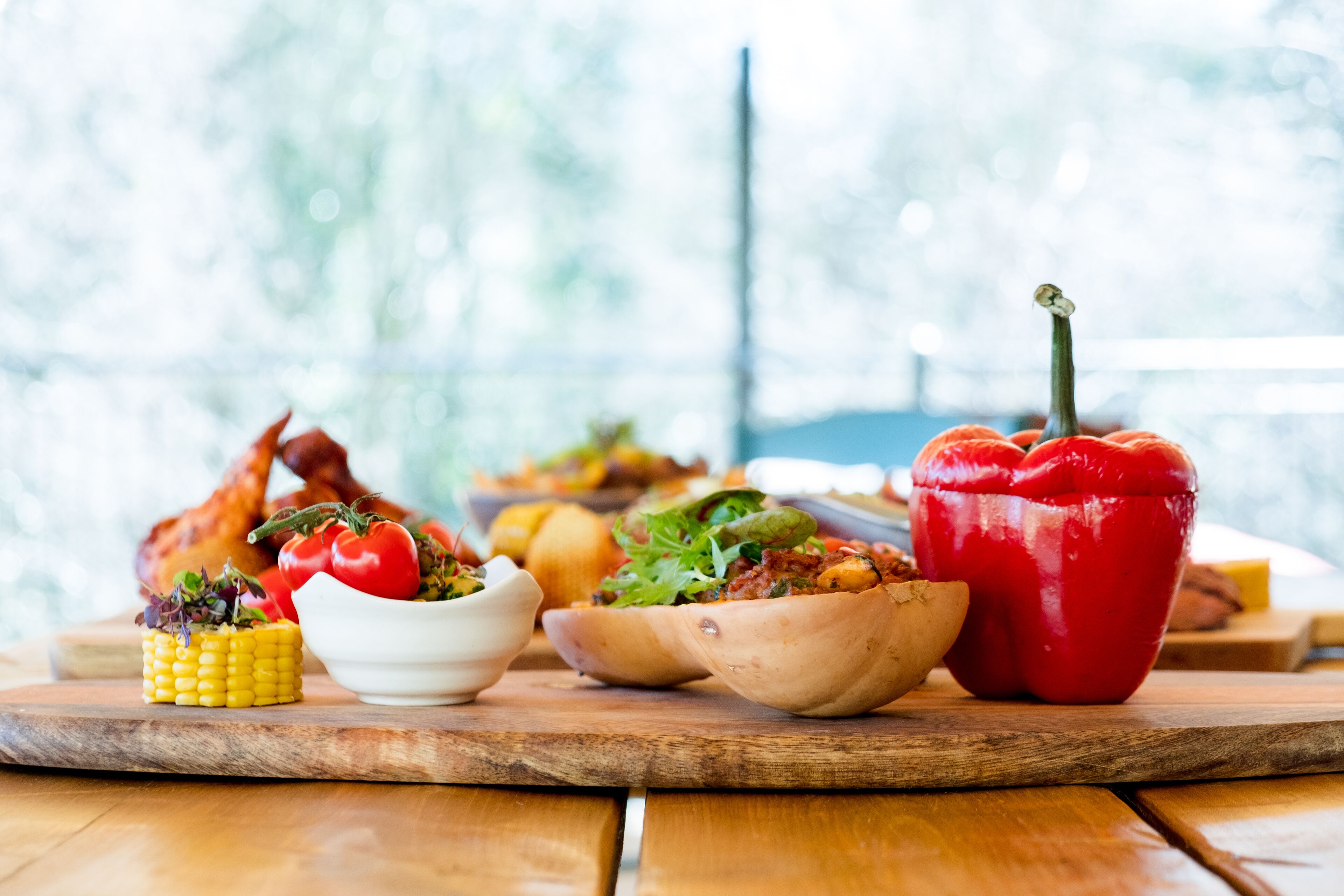 A wooden table with various dishes including corn, tomatoes, and a red bell pepper.