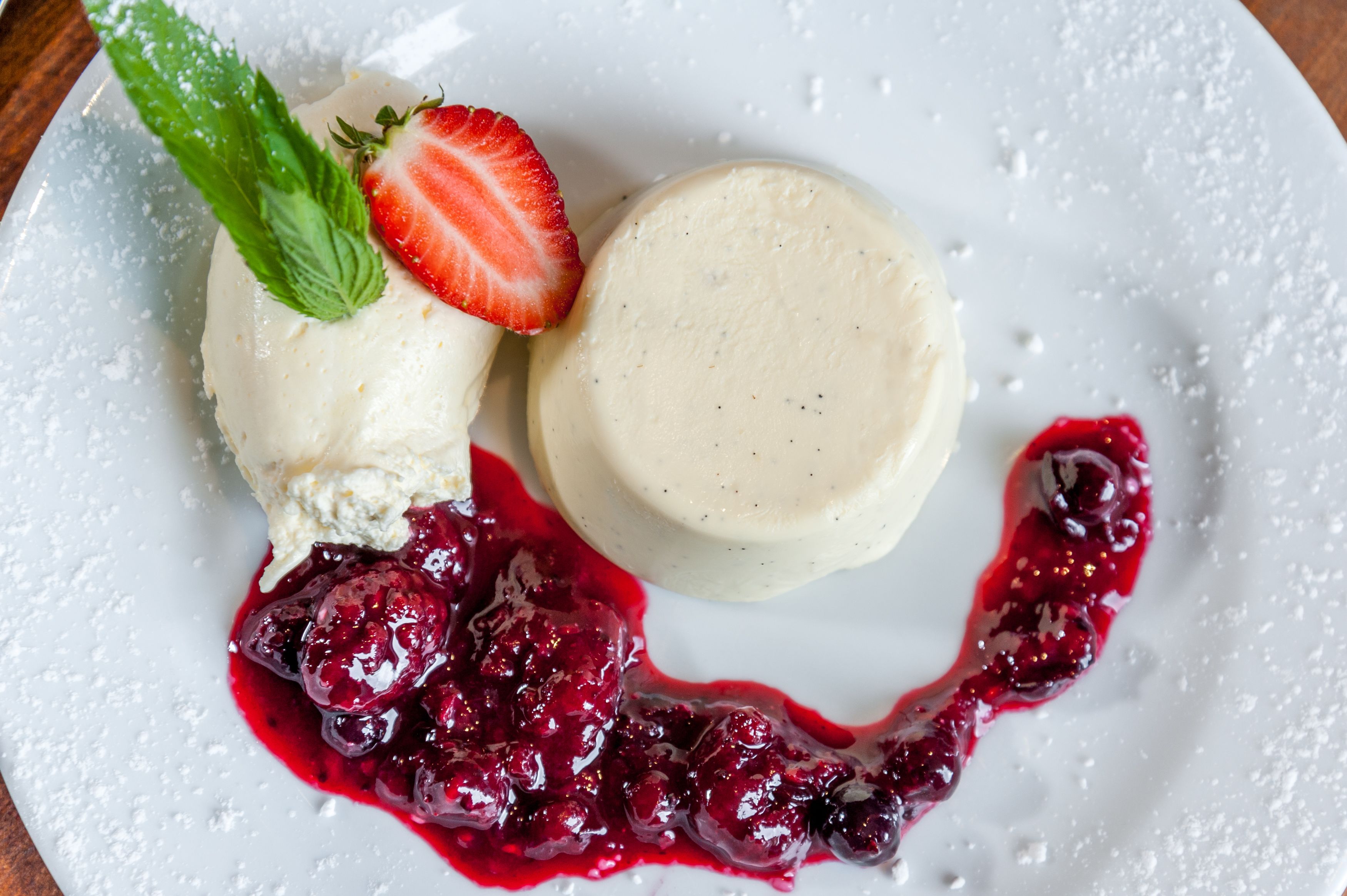 Un plato de postre con una bola de helado de vainilla, una porción de panna cotta y un coulis de frutos rojos.