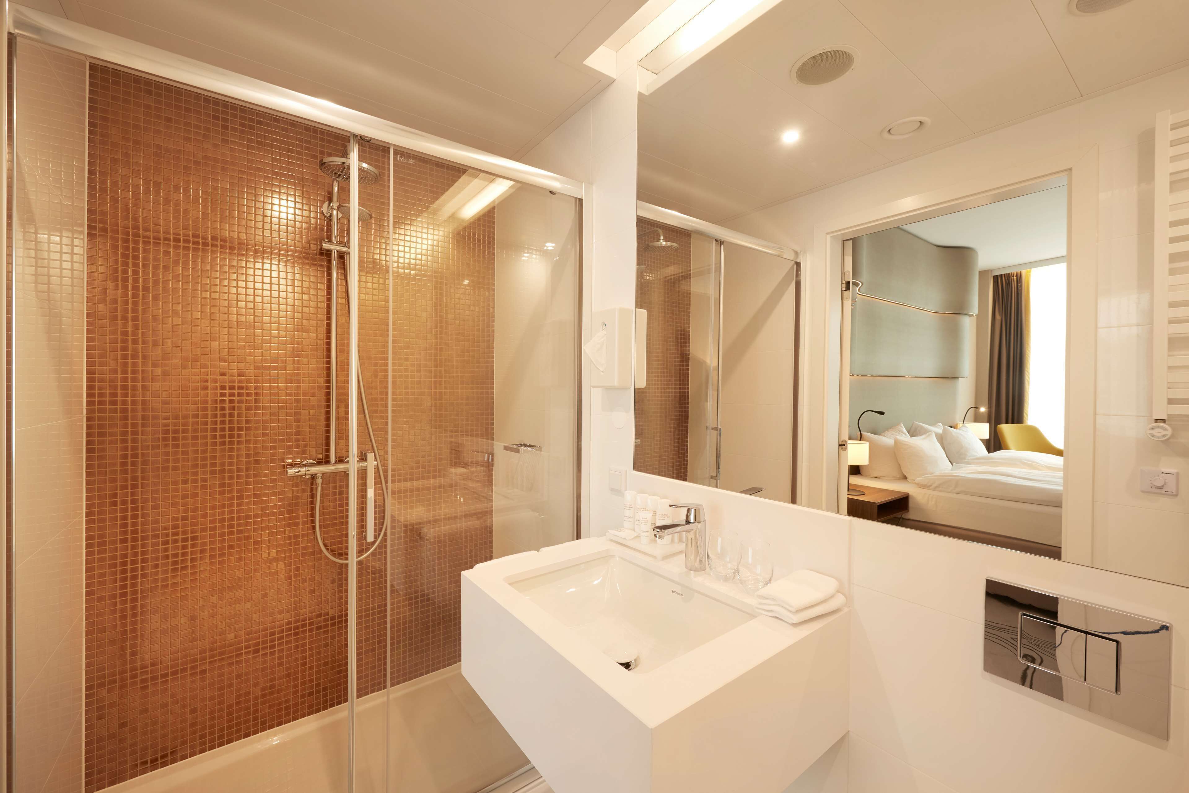 A modern bathroom with a glass-enclosed shower, a white sink, and a large mirror reflecting a bedroom.