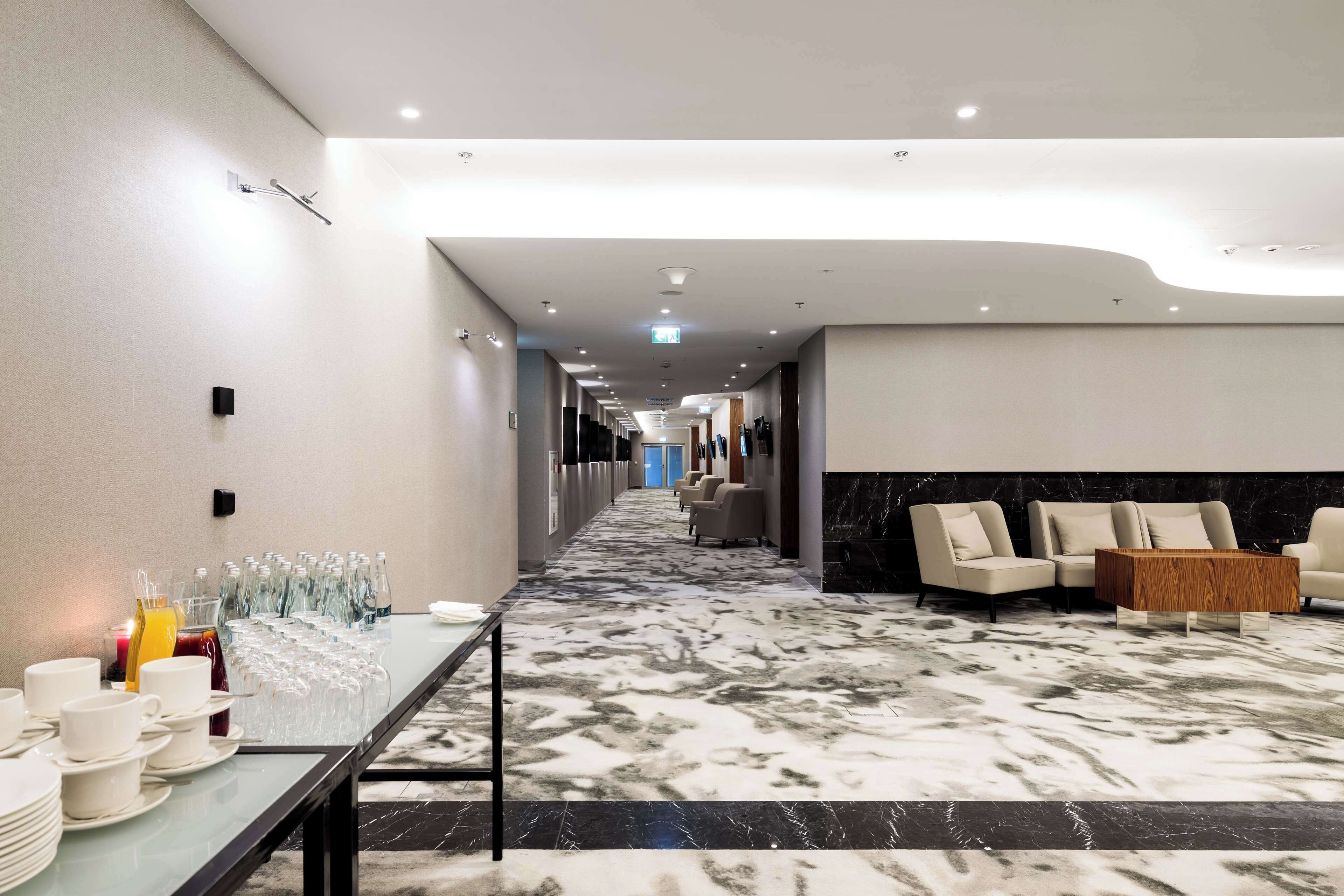 Eine Hotelhalle mit Marmorboden, einer Sitzecke mit Sofas, einem langen Tisch mit Frühstücksangeboten und einem modernen Deckenstil.