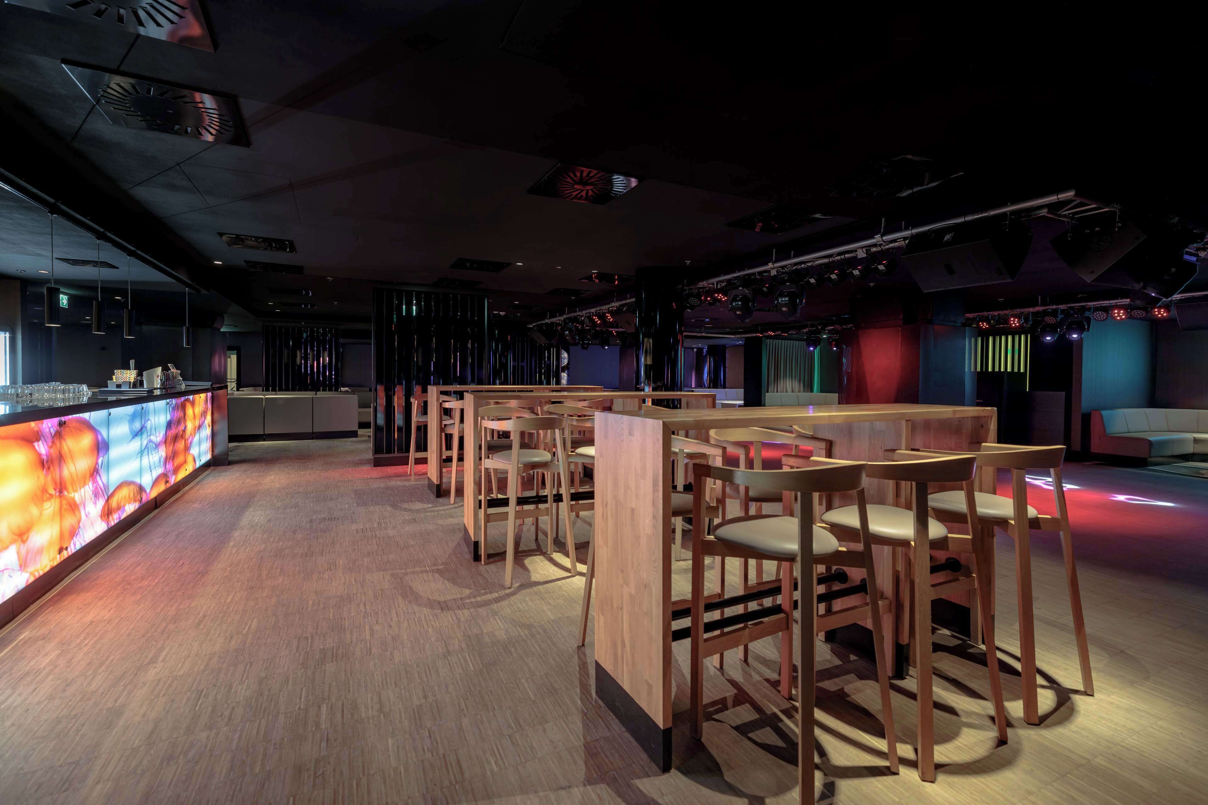 Zona de bar moderna con mesas y sillas de madera, una barra con pantalla digital y un escenario con equipo de iluminación.