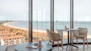 Ein modernes Hotelzimmer mit großem Fenster und Blick auf Strand und Meer. Zwei Sessel und ein kleiner Tisch mit Kerze und Buch.