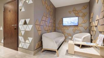 A modern hotel room with a wooden door, a beige sofa, a wooden coffee table, and a TV mounted on the wall.
