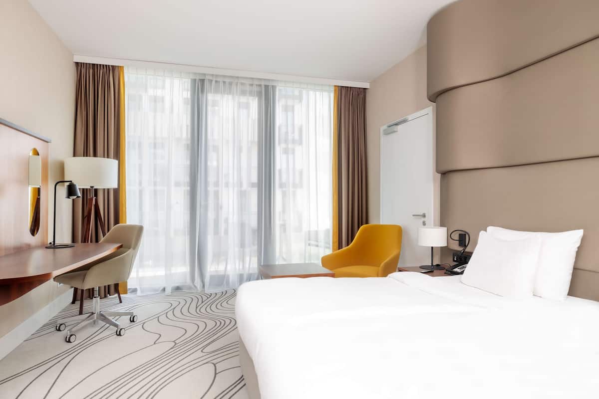 A hotel room with a large bed, a desk with a chair, a yellow armchair, a large window with curtains, and a modern design.