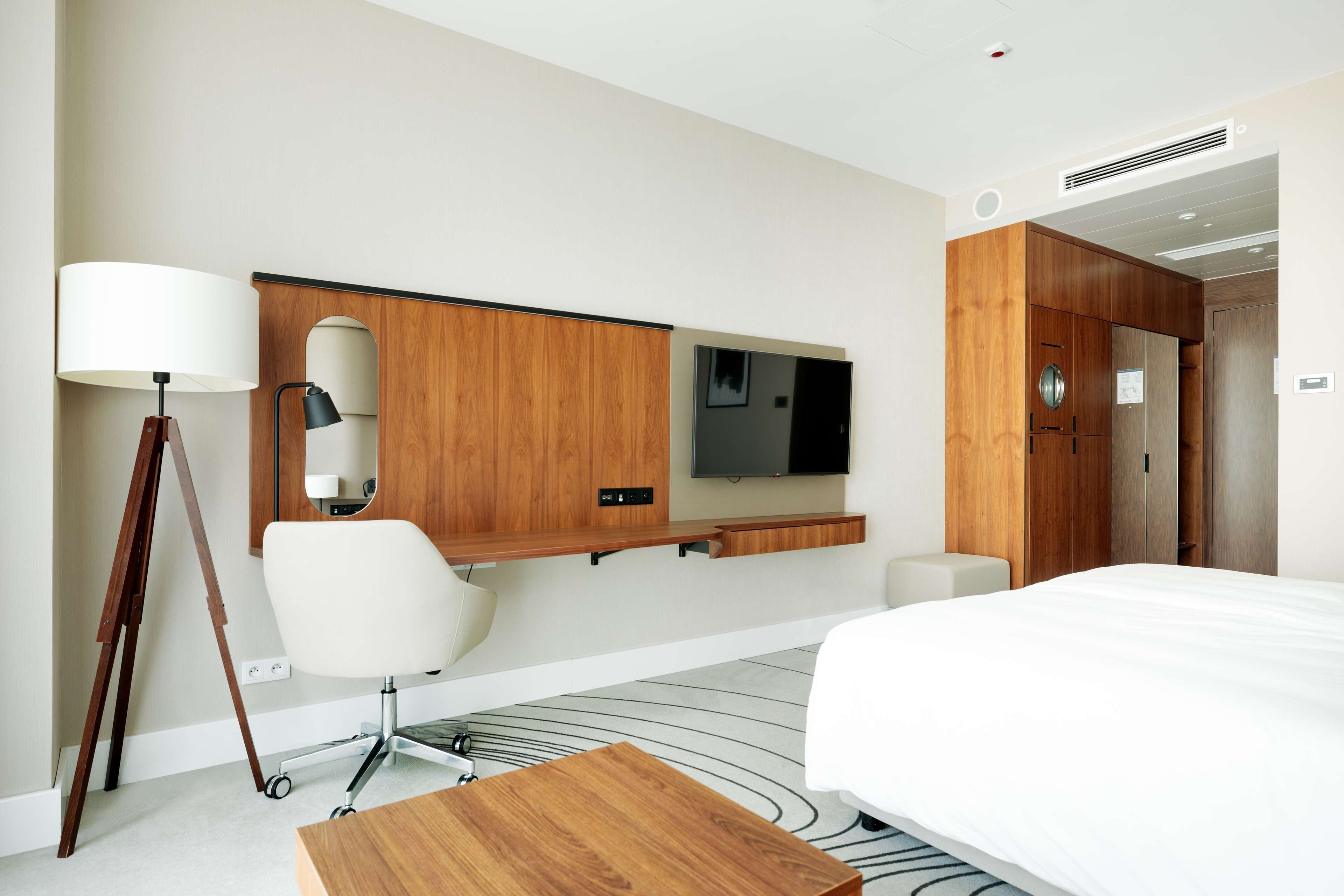 A hotel room with a bed, a desk with a chair, a standing lamp, a flat-screen TV, and a wooden wardrobe.