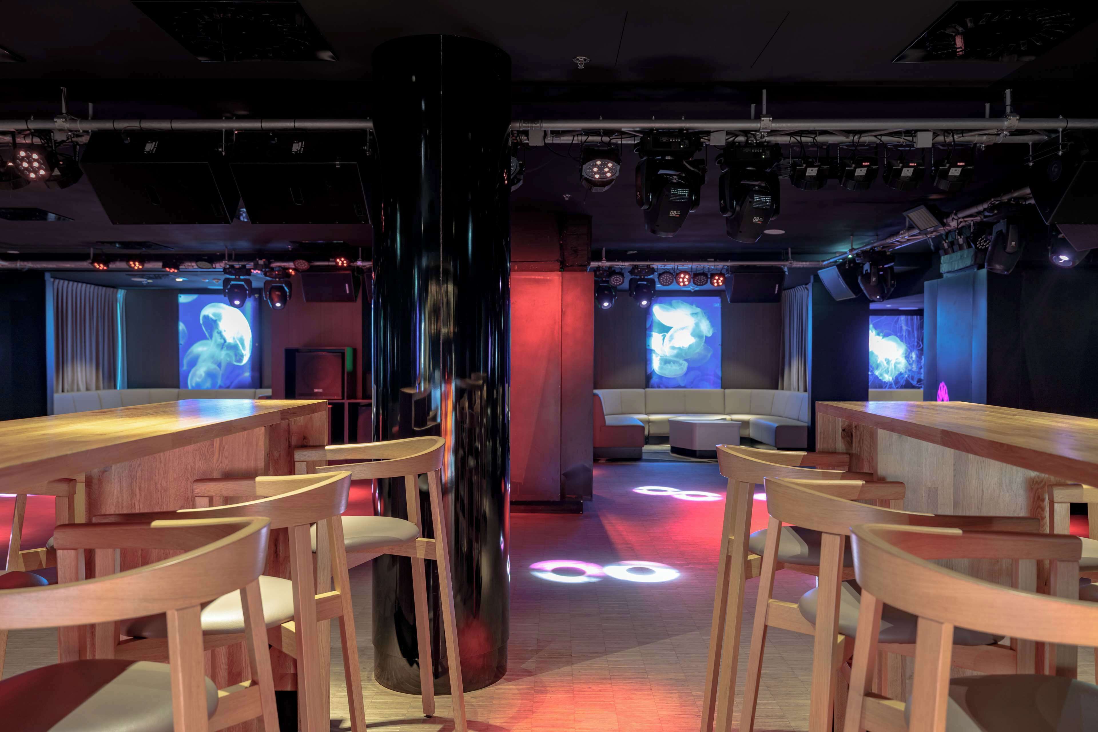 A modern bar area with wooden tables and chairs, large screens displaying visuals, and overhead lighting.
