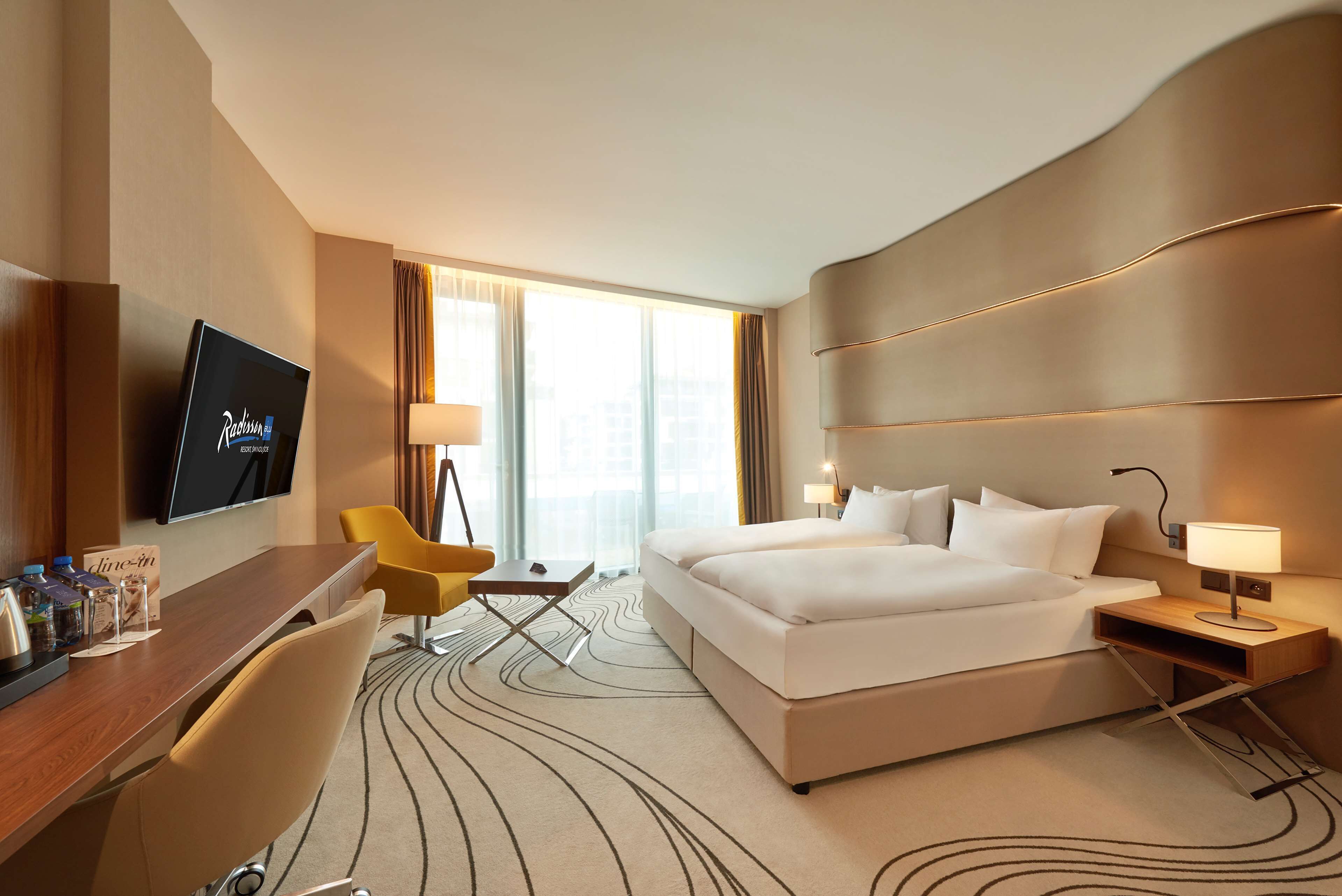 Habitación de hotel con una cama grande, un escritorio con televisor de pantalla plana, una silla, una lámpara de escritorio y una ventana con cortinas.