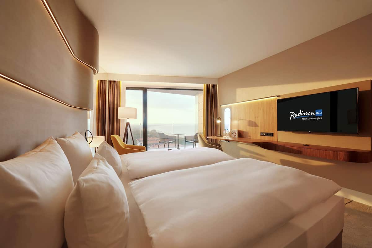 A hotel room with two beds, a TV, a desk, and a view of the outdoors.