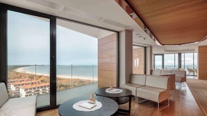 Ein modernes Zimmer mit großem Balkon, einer Couch, zwei runden Tischen und Blick auf Strand und Häuser.