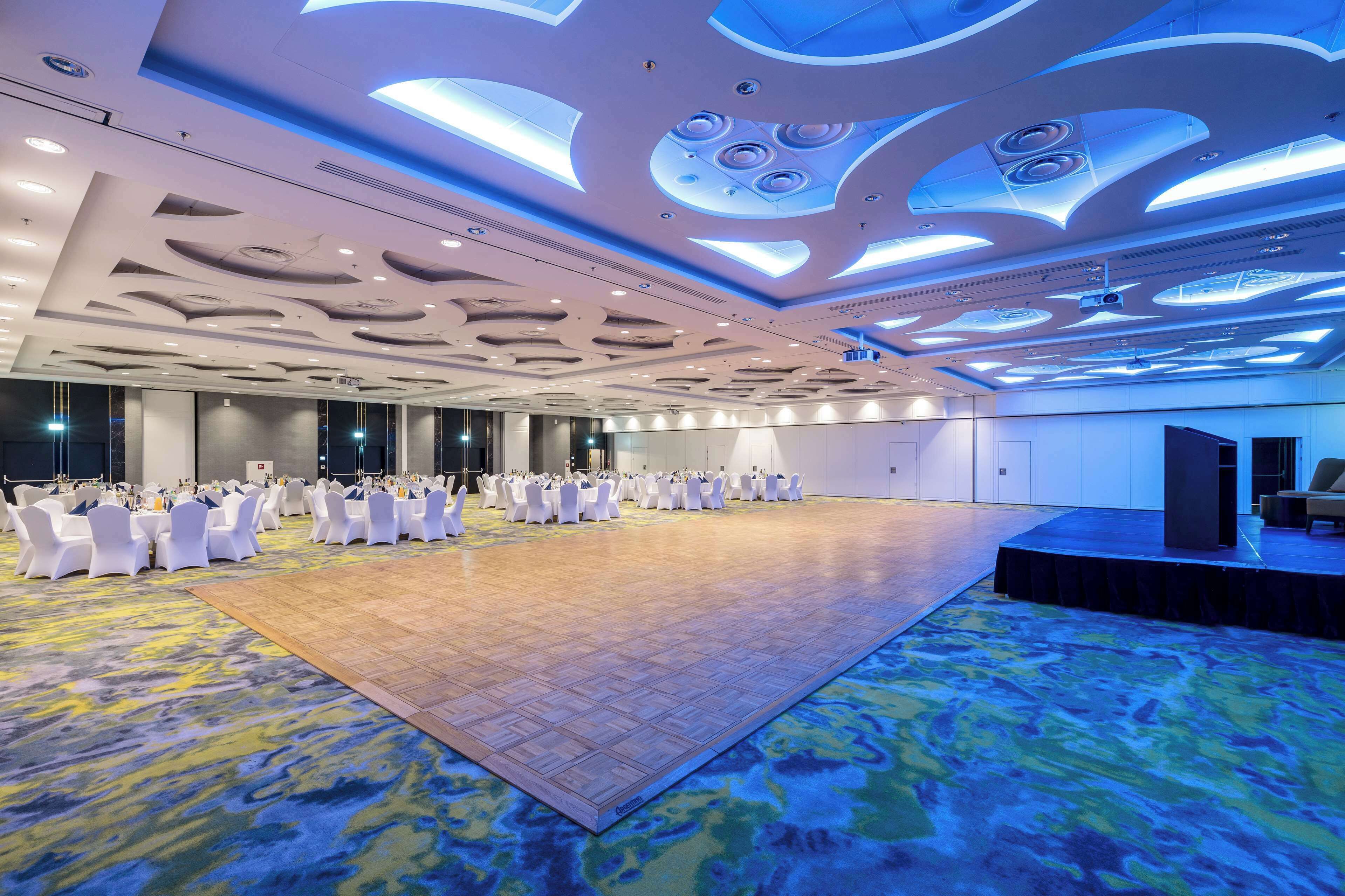 A spacious conference room with white chairs and tables, a stage, and a patterned carpet.
