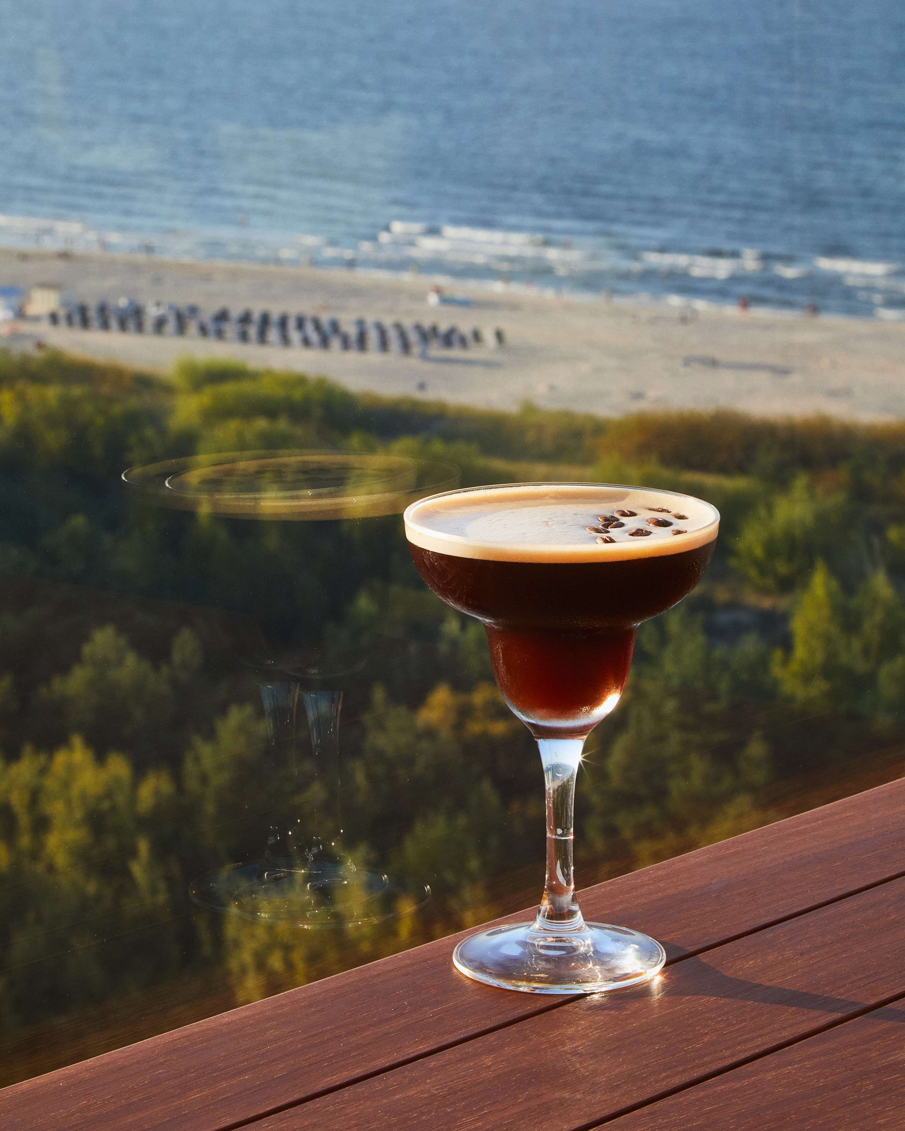 A martini glass with a frothy top on a wooden deck, overlooking a beach and ocean.