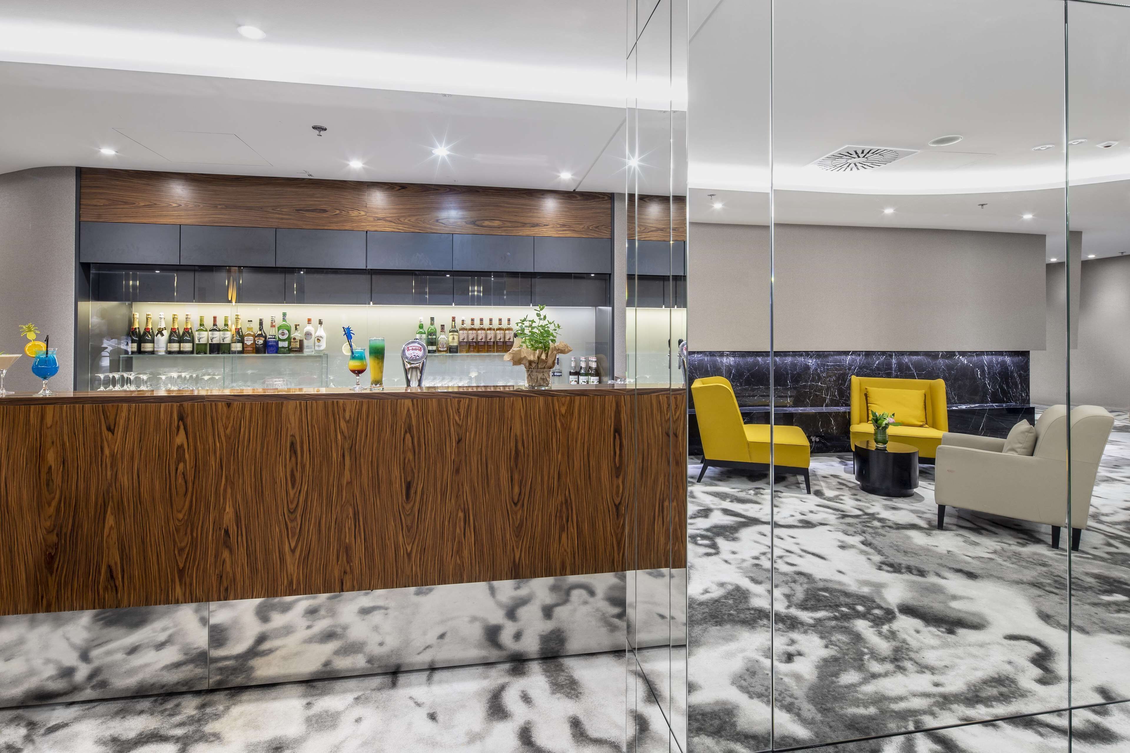 A modern bar area with a wooden counter, a variety of bottles, and a seating area with yellow chairs and a white sofa.