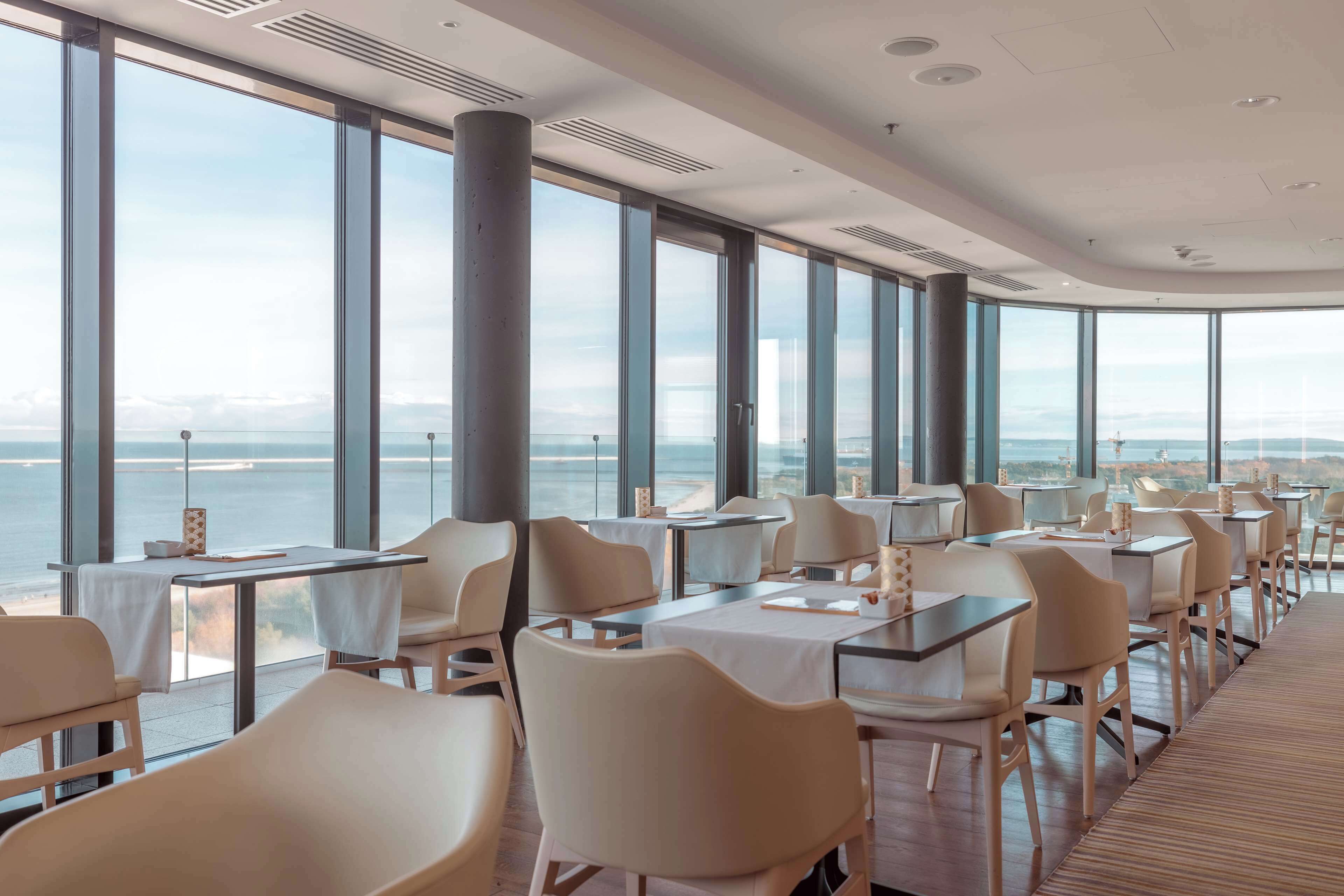 A modern dining area with large windows, tables, and chairs, overlooking a body of water.
