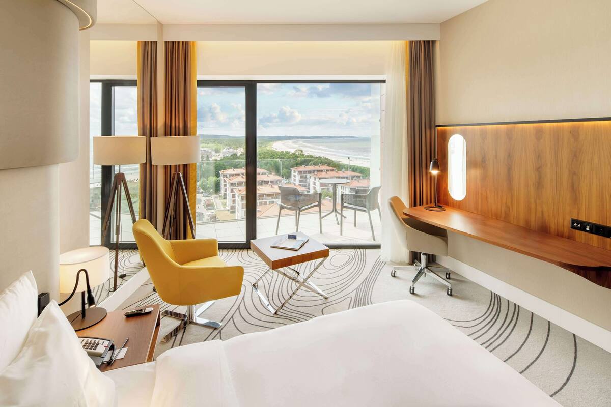 A hotel room with a large bed, a desk with a lamp, a chair, and a view of buildings and a body of water.