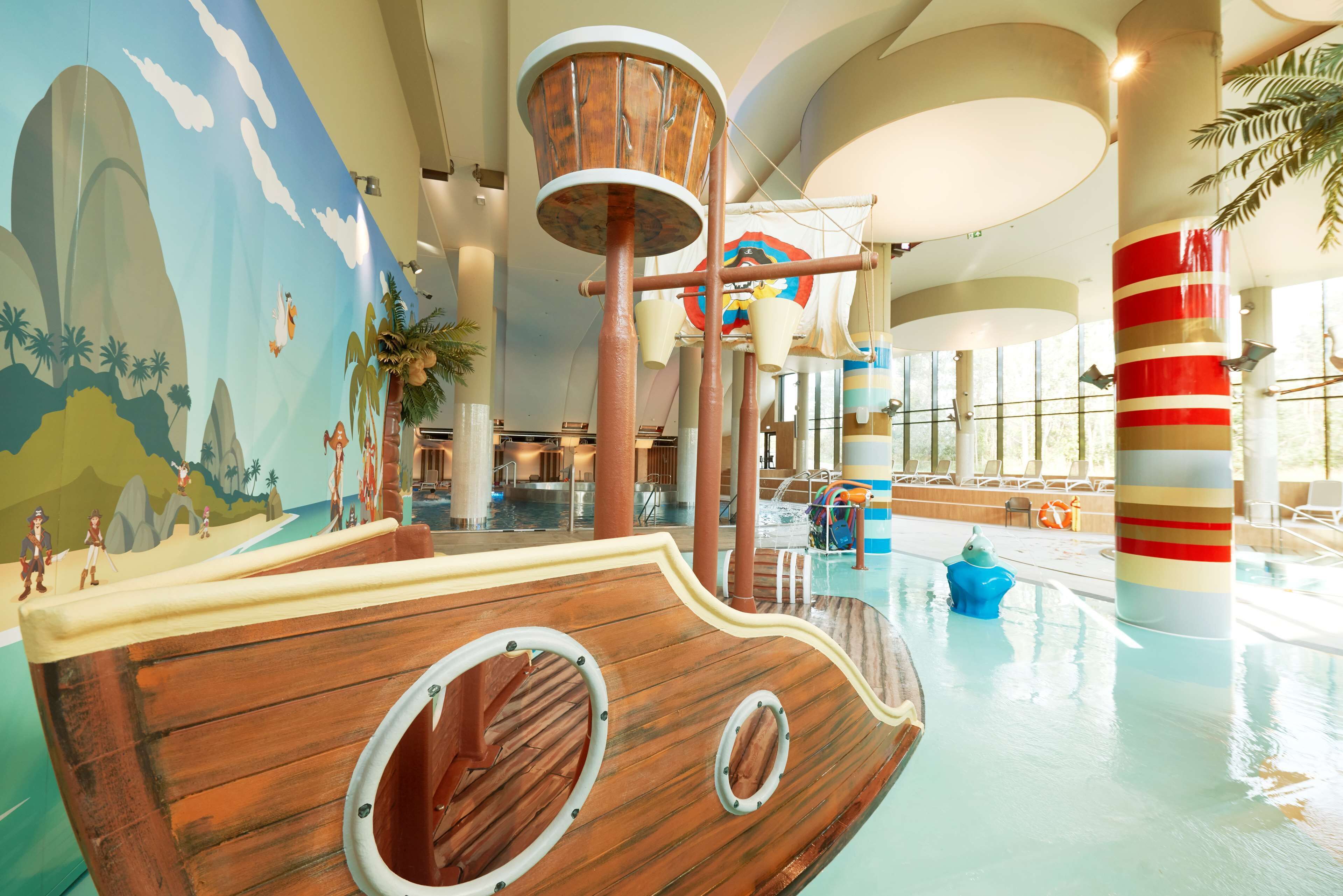 Un amplio espacio de juegos cubierto con una estructura temática de barco, una piscina y un mural con una playa.