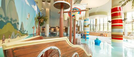 Un amplio espacio de juegos cubierto con una estructura temática de barco, una piscina y un mural con una playa.