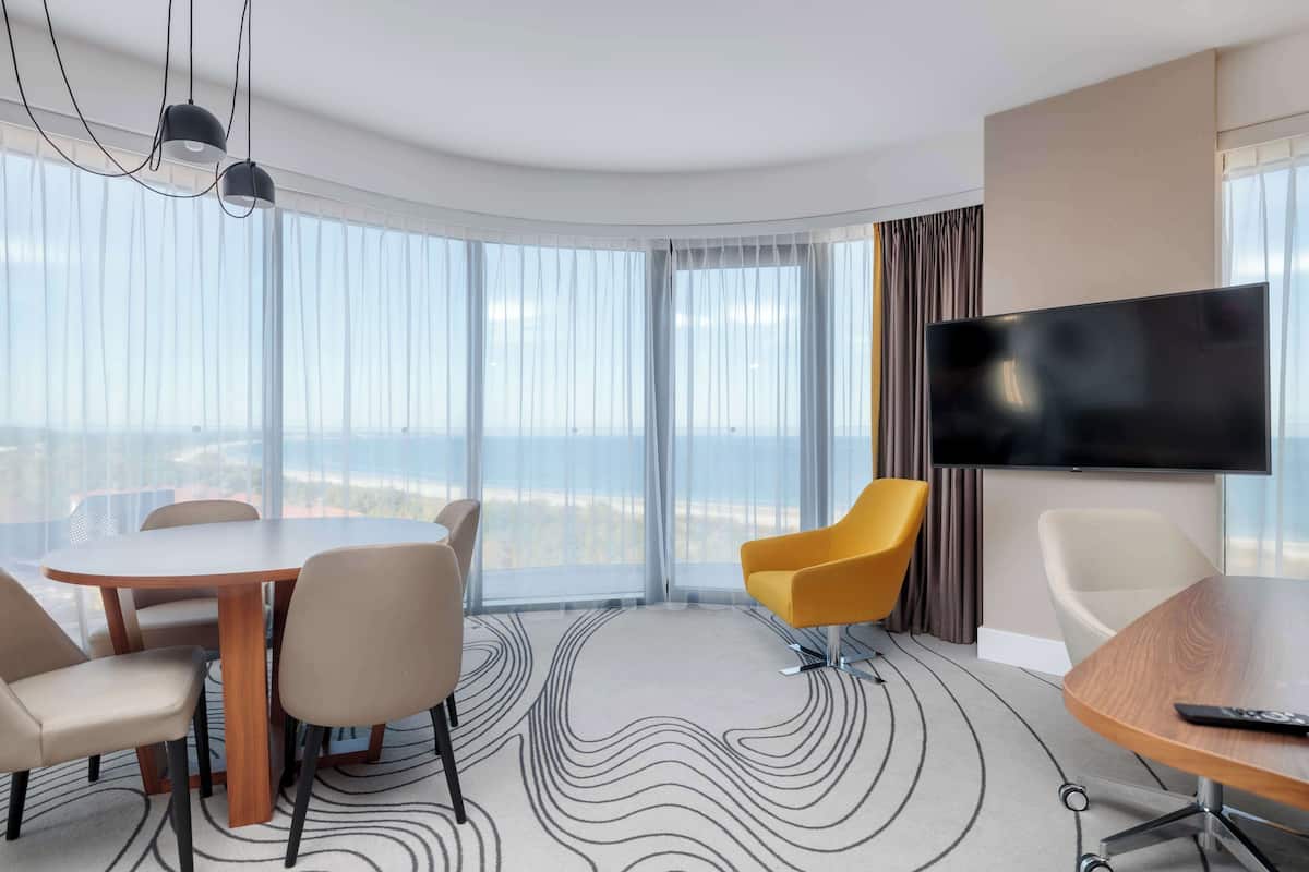 A hotel room with a round table, four chairs, a TV, and a large window with curtains.