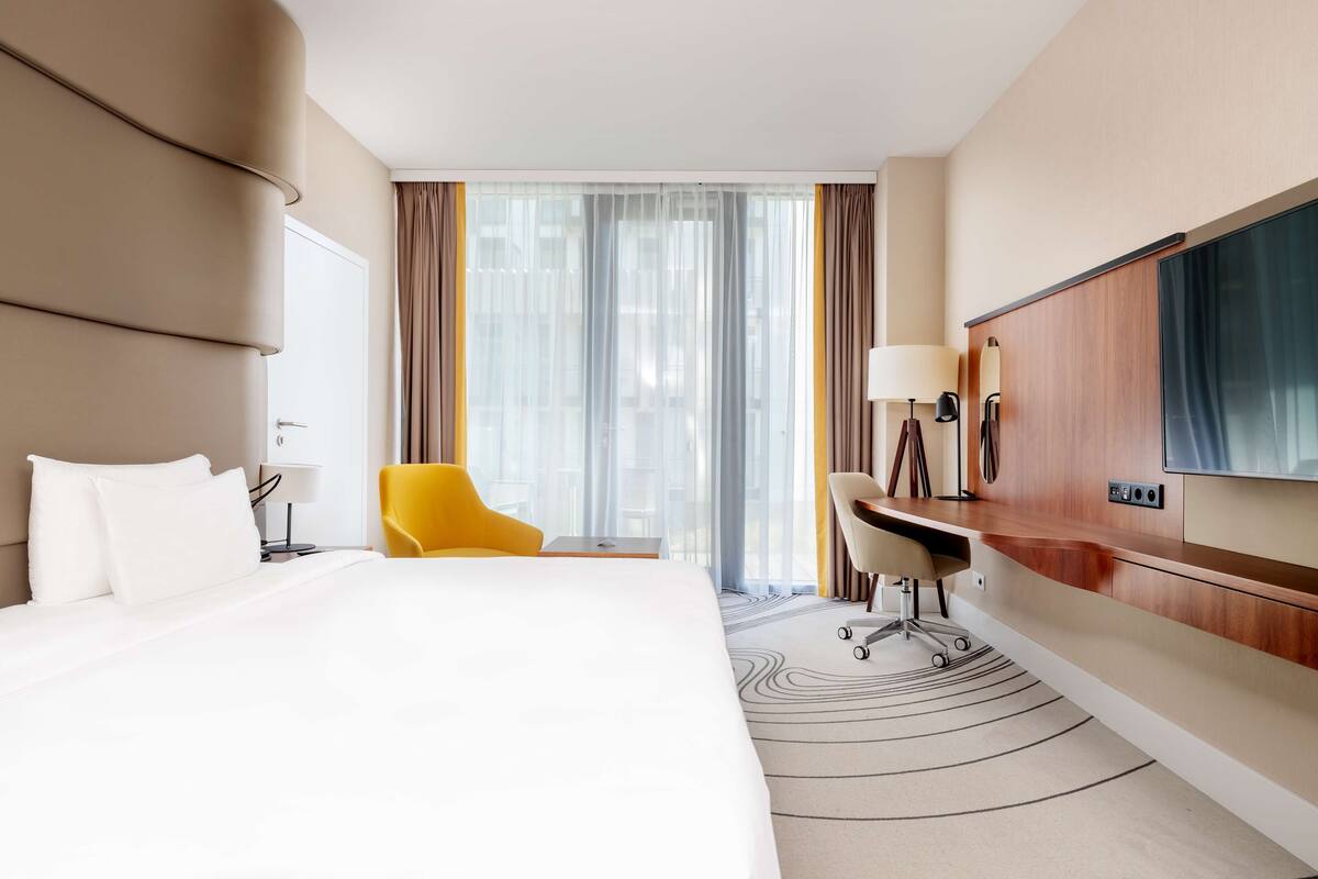 A hotel room with a large bed, a desk, a chair, a TV, and a view of the city through the window.