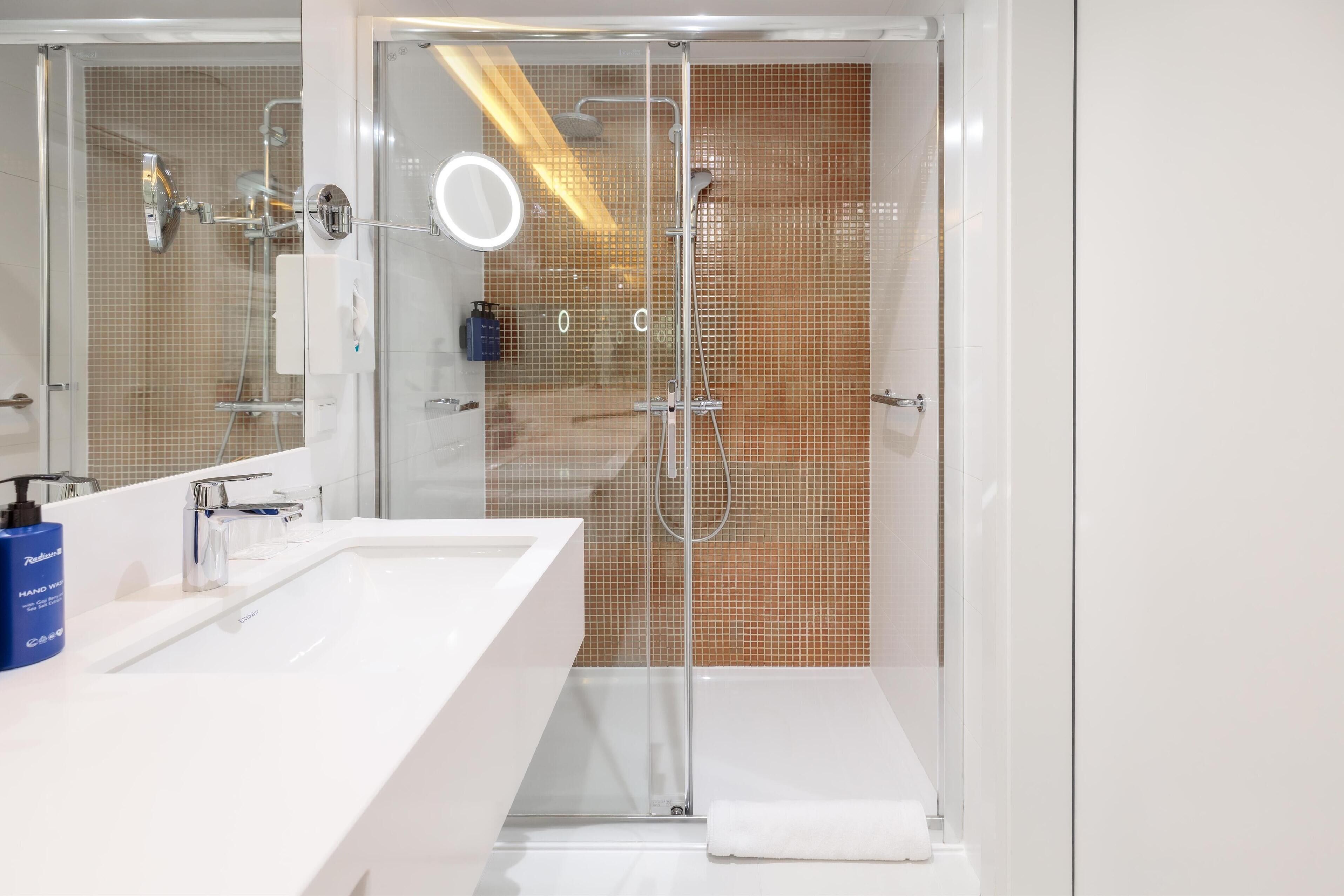 Un baño moderno con un lavamanos blanco grande, un box de ducha de vidrio y un espejo con luz circular.