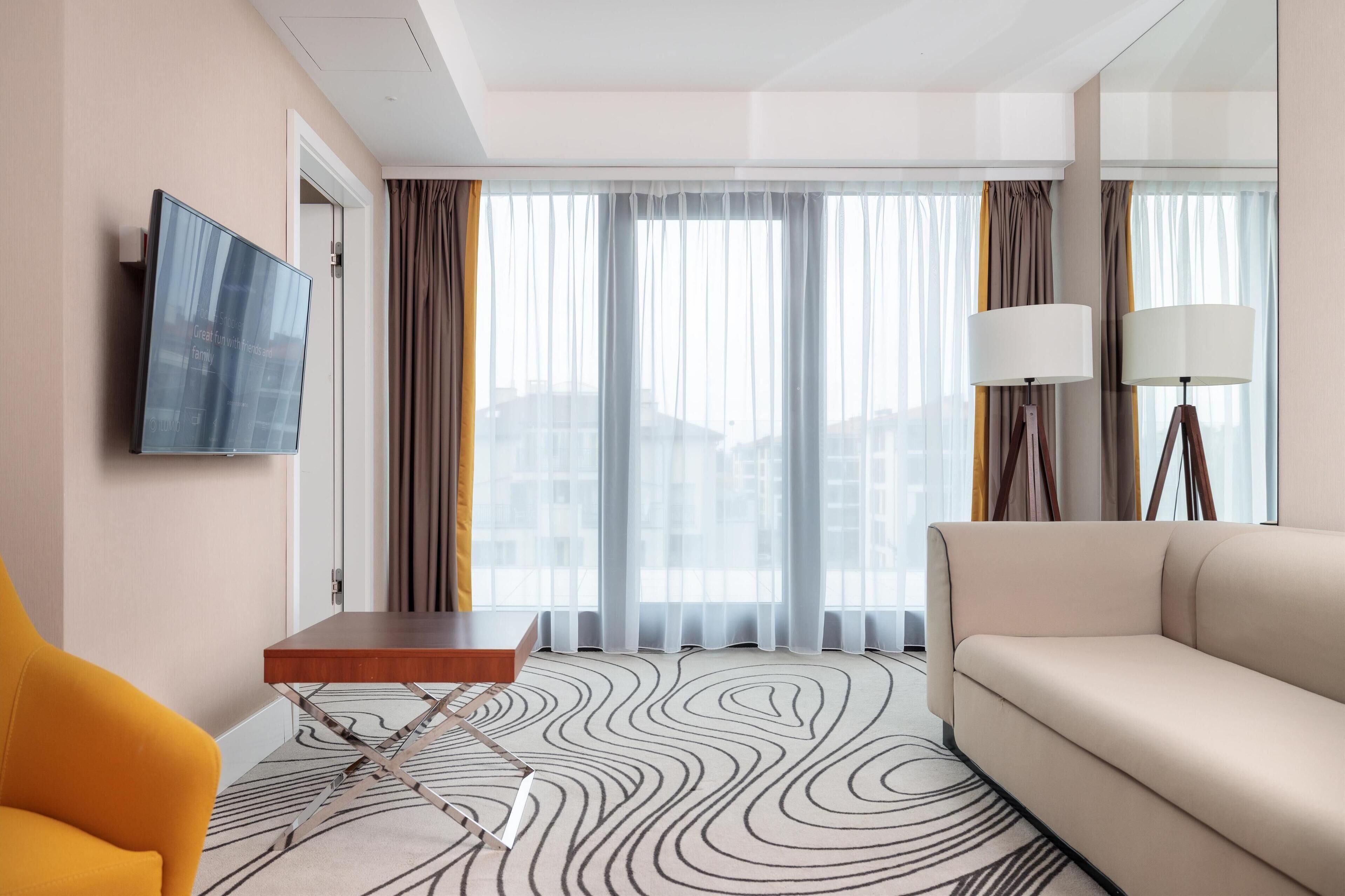 Una habitación de hotel con sofá, una mesita, televisor montado en la pared y ventana con cortinas.