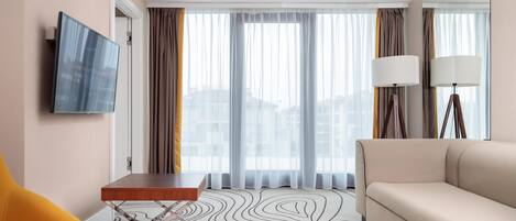Una habitación de hotel con sofá, una mesita, televisor montado en la pared y ventana con cortinas.