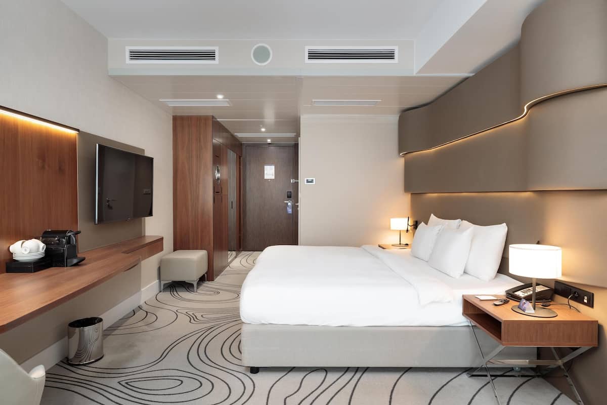A hotel room with a large bed, a bedside table with a lamp, a TV mounted on the wall, and a wooden desk with a coffee maker.