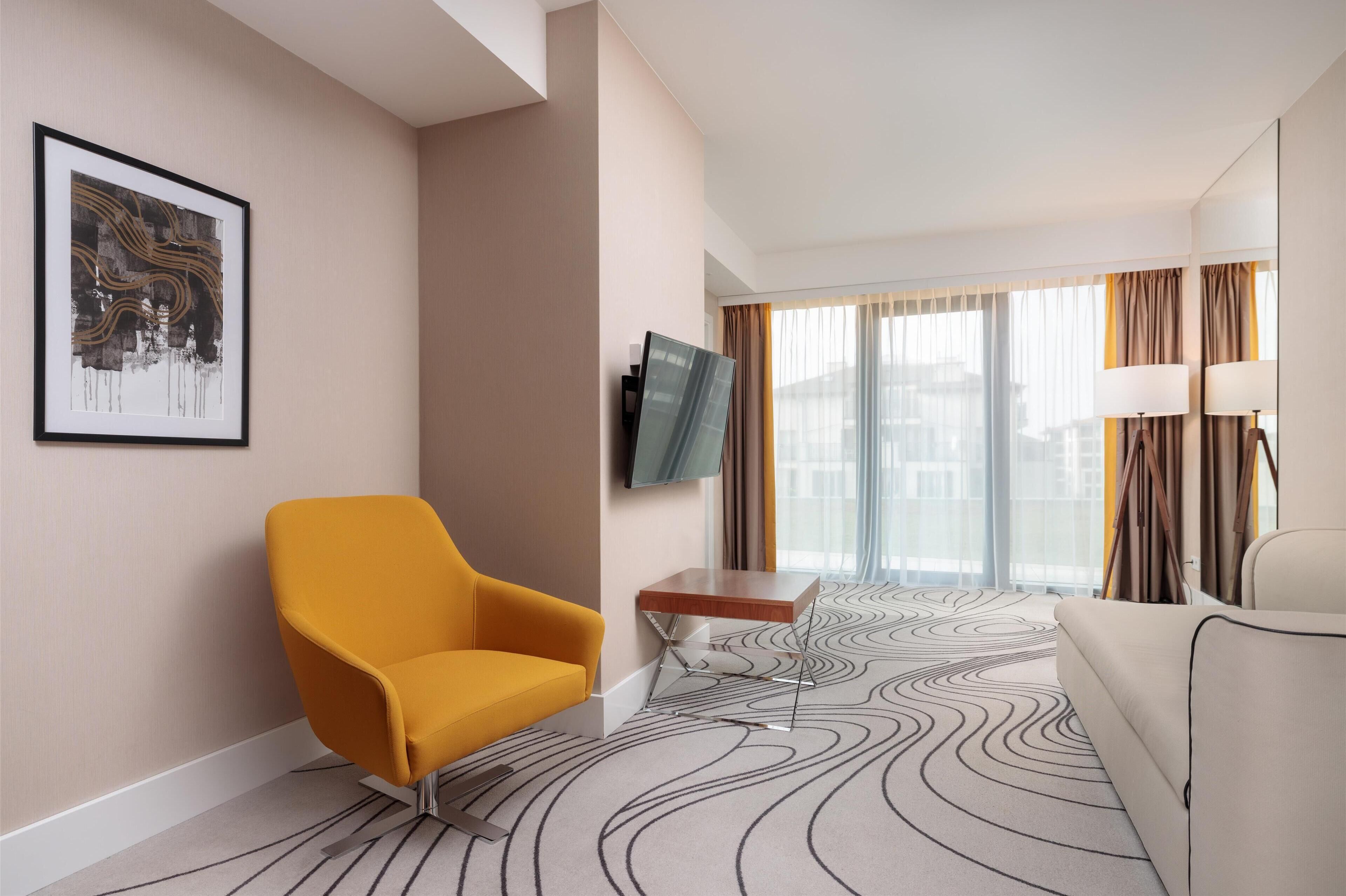A modern hotel room with a large window, a flat-screen TV, a yellow armchair, a white sofa, and a patterned carpet.