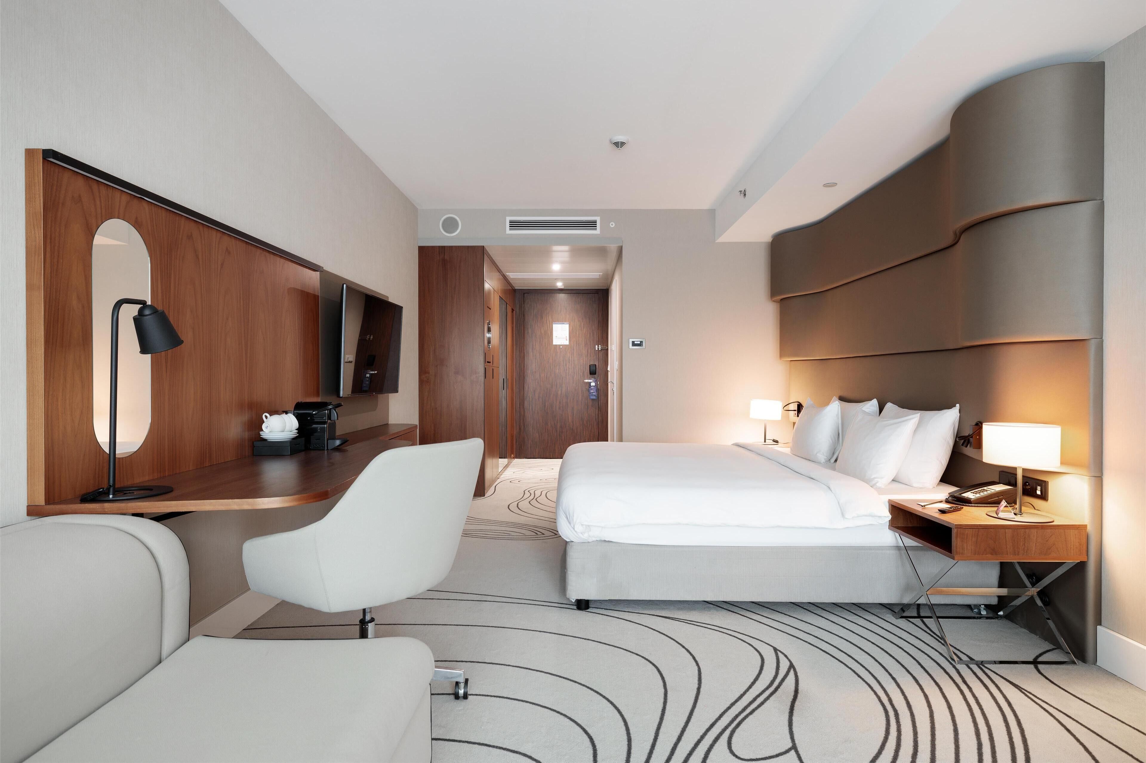 A hotel room with a bed, a desk with a lamp, a chair, a TV, and a mirror.