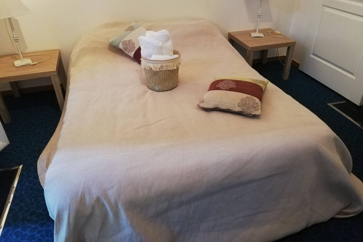 Een net opgemaakt bed met een beige sprei, twee kussens, een klein mandje met een handdoek en twee bedlampjes op houten nachtkastjes.