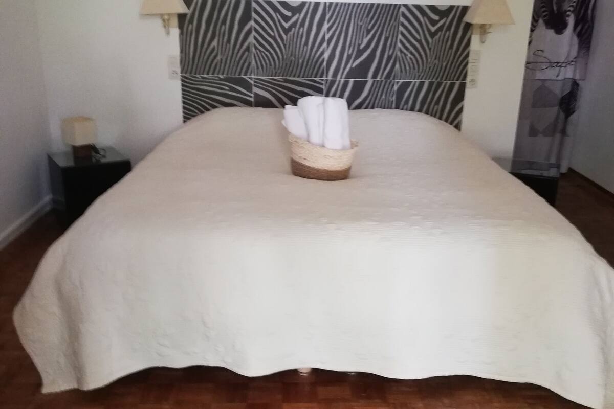 Een net opgemaakt bed met een hoofdeinde met zebrapatroon, een nachtkastje met een lamp, en een wand met behang in zebraprint.