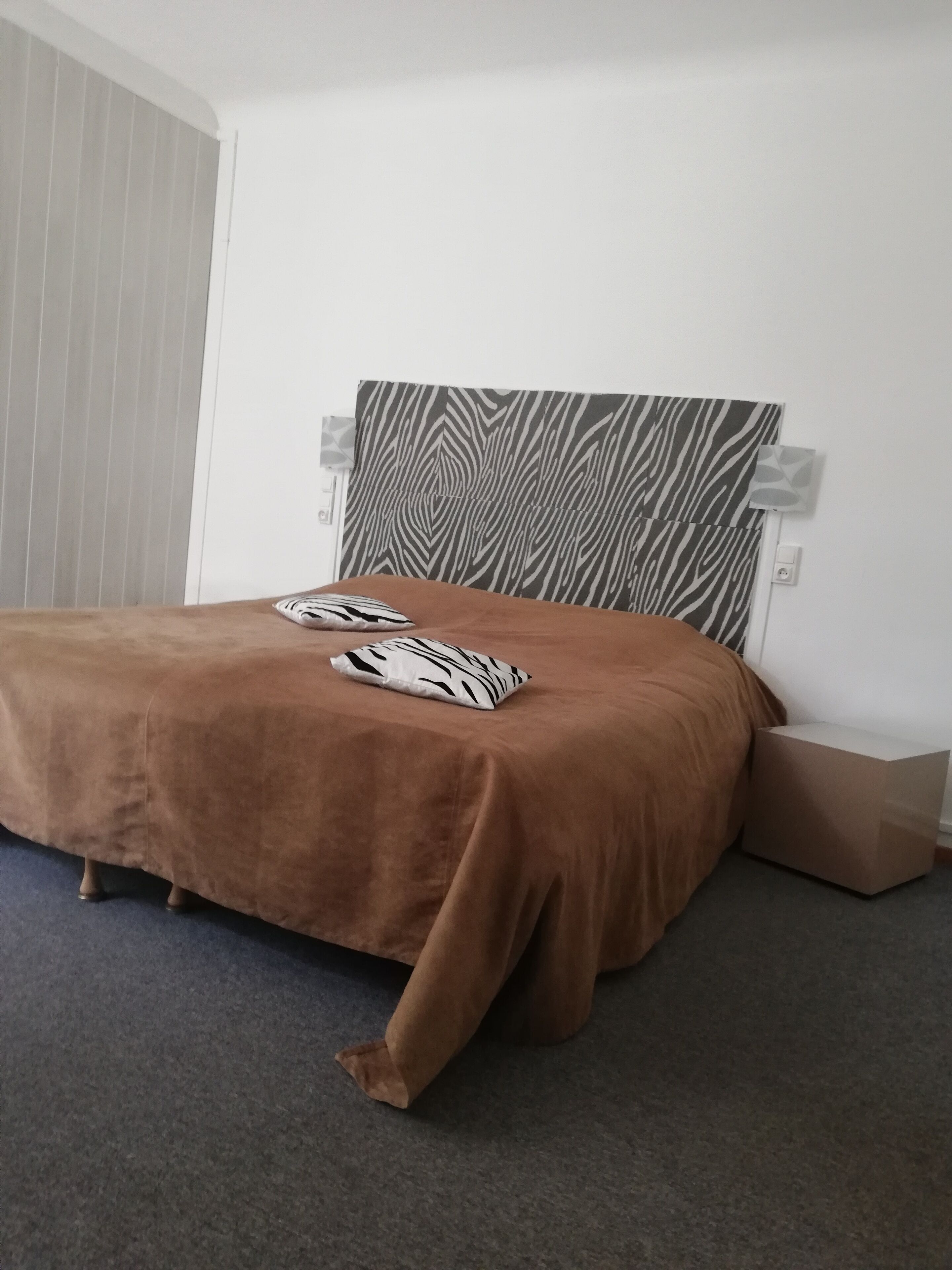 Una camera da letto con una coperta marrone, cuscini a motivi zebrati e un poggiatesta a righe zebrate. Si nota una parete bianca con un interruttore della luce e un piccolo oggetto bianco sopra.