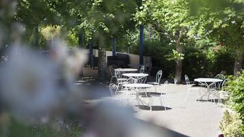 An outdoor patio with white metal tables and chairs, surrounded by trees and greenery.