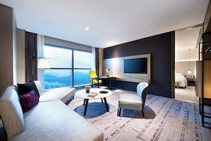 A modern hotel room with a large window, a flat-screen TV, a sofa, a coffee table, and a chair.
