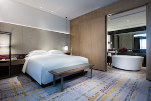 A modern hotel room with a large bed, a bench, a bathroom with a bathtub, and a large mirror.