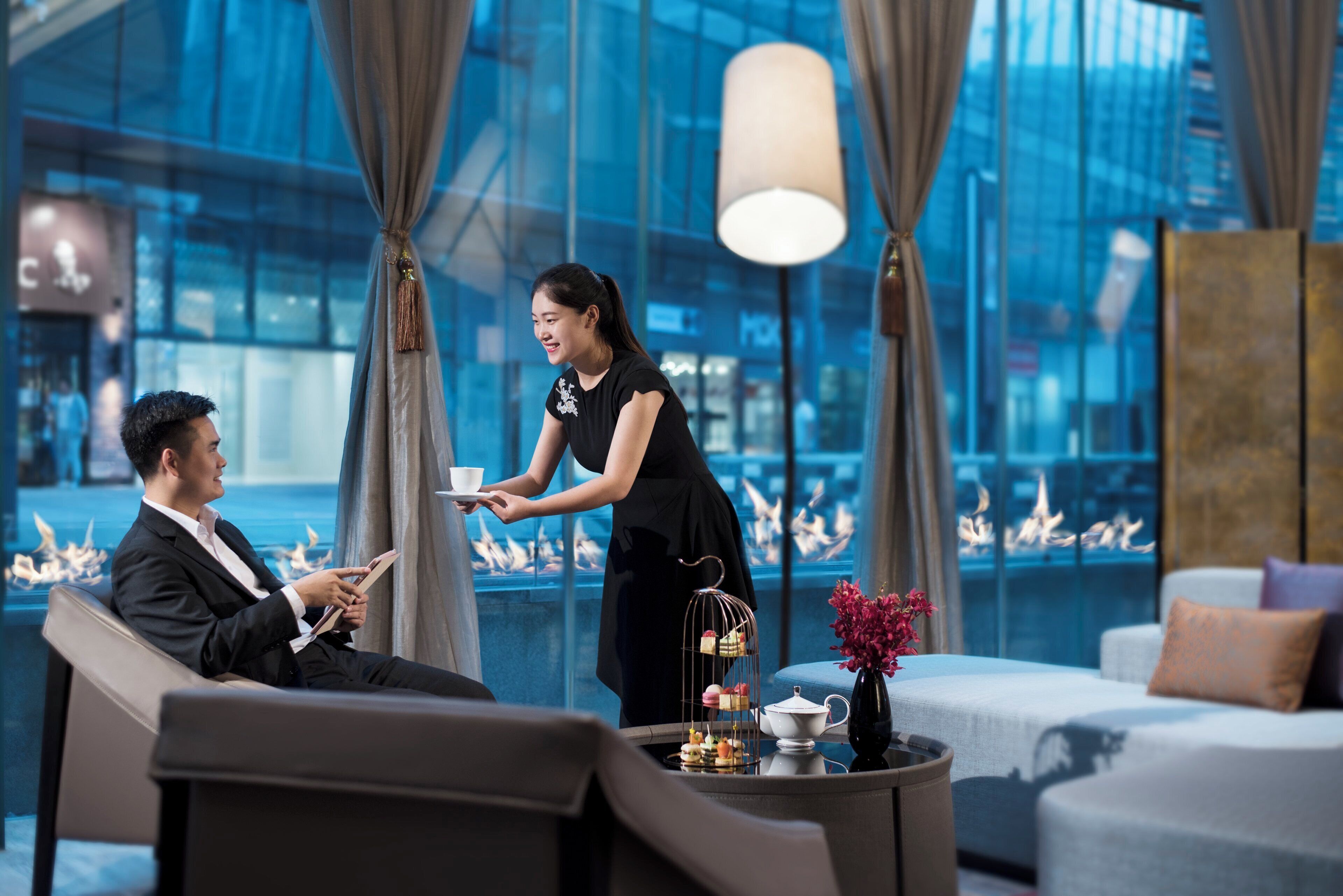 A modern hotel lobby with a seating area, a reception desk, and a large window with a city view.