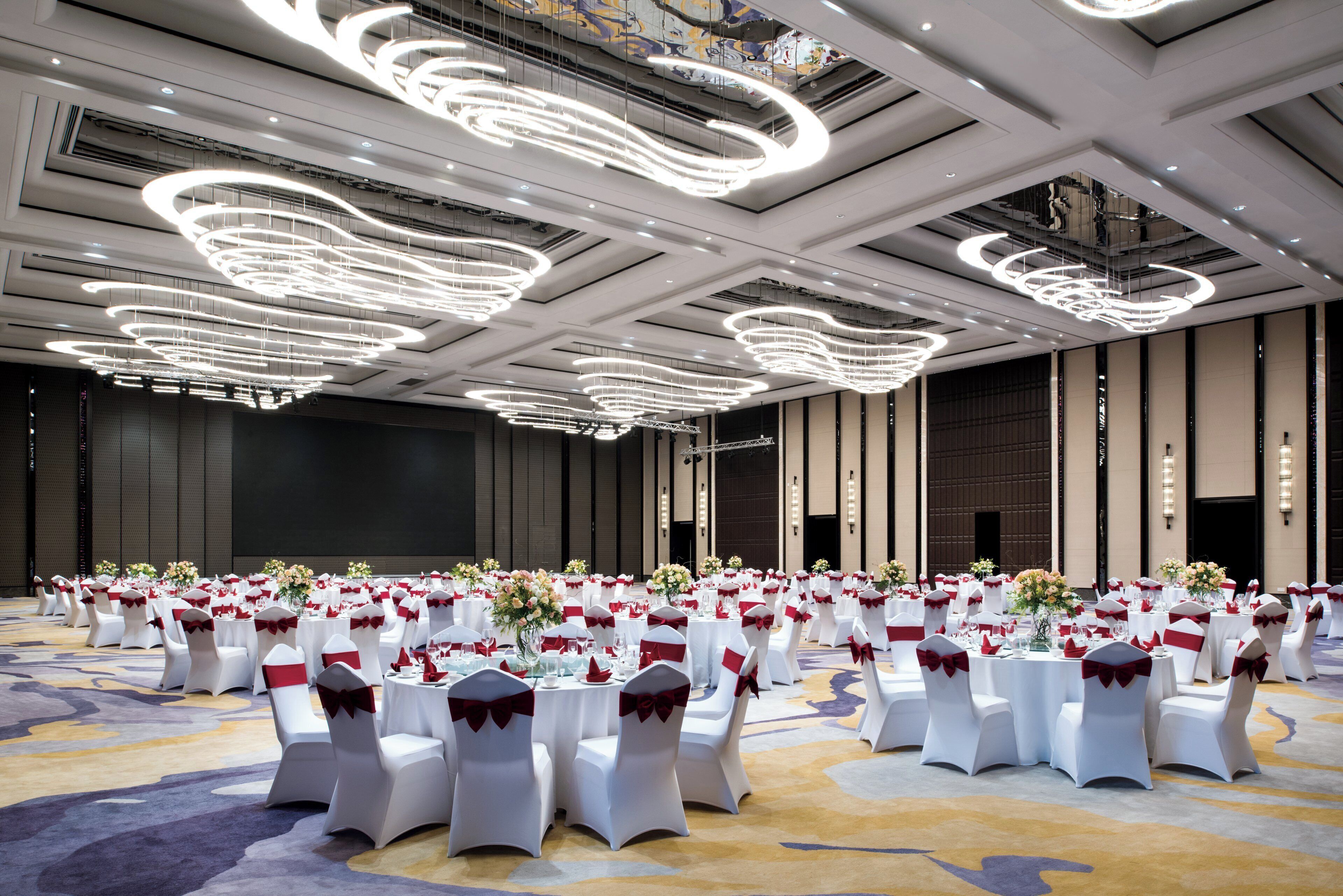 A banquet hall with round tables, white chairs with red sashes, and modern circular ceiling lights.